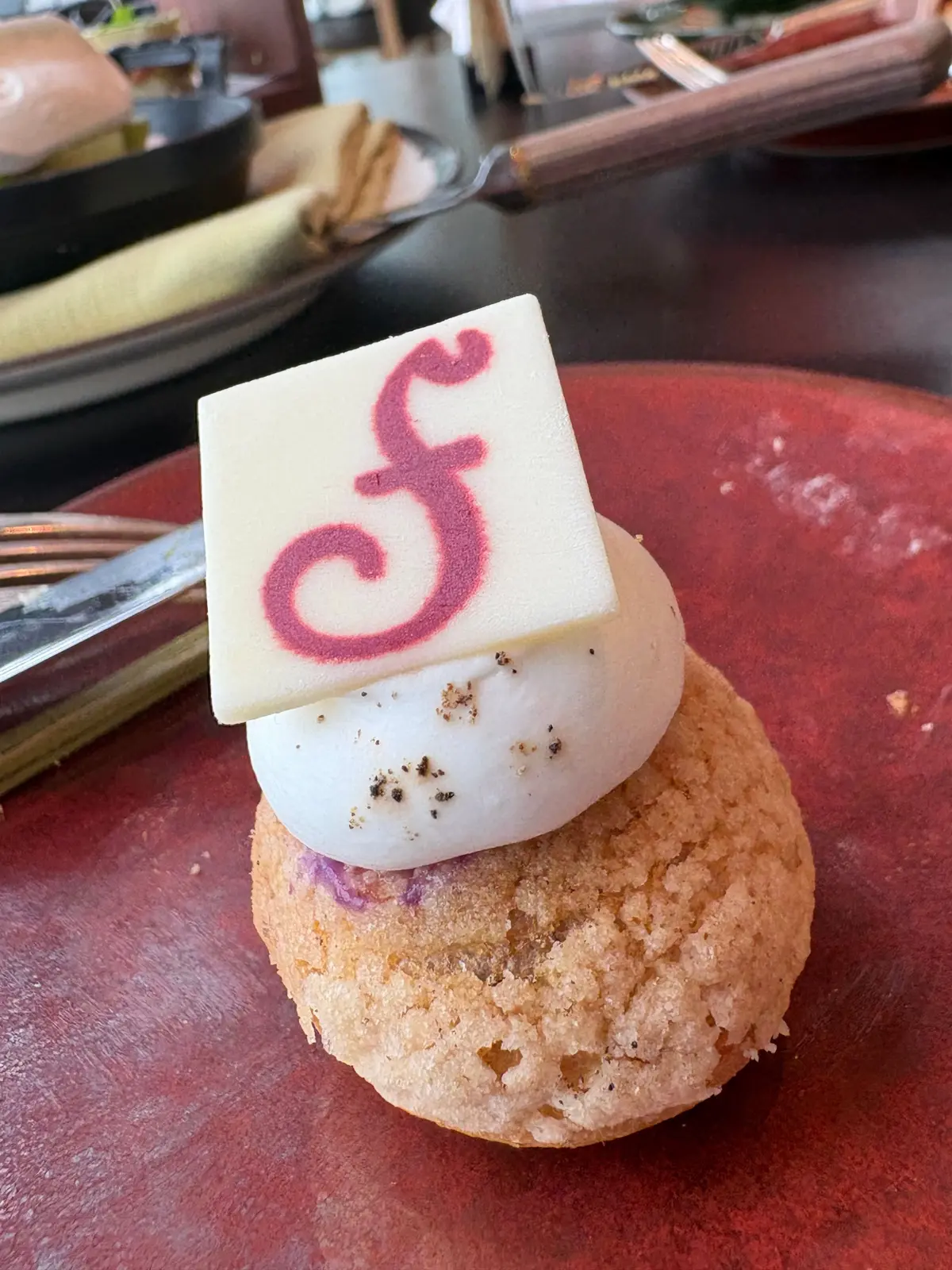 赤茶色の円盤の上に、丸い焼き菓子、白いムース、四角い砂糖菓子が積み重ねられている。砂糖菓子には赤い文字で「F」の模様が描かれている。