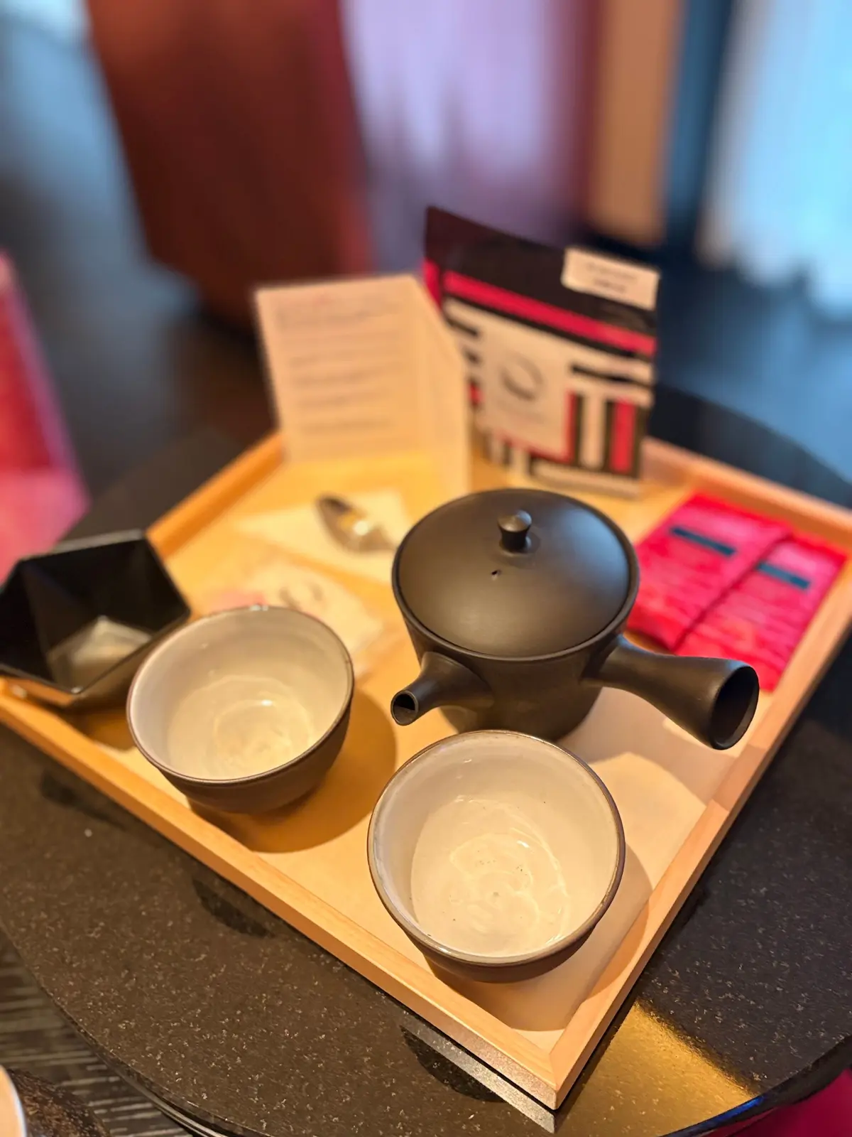 木製のトレイに、茶器と茶葉、説明書が置かれている。茶器は黒色で、2つの白い湯呑みと、黒い小皿が添えられている。背景には、ピンクと青のカーテンが見える。