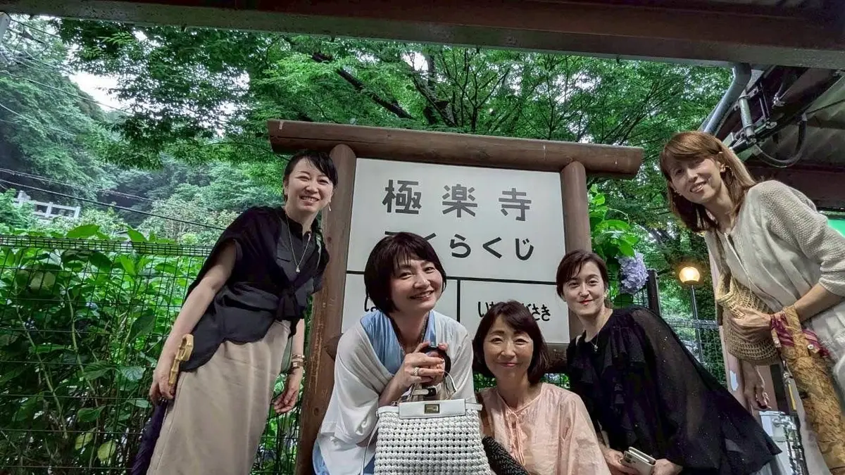 桜茶店と書かれた看板の前で、５人の女性が笑顔で並んでいる。手前から右に、白の編みかごバッグを持つ女性、ピンクの服の女性、黒の服の女性、左に黒の服の女性、右にベージュの服の女性が立っている。背景には緑の木々が広がる。