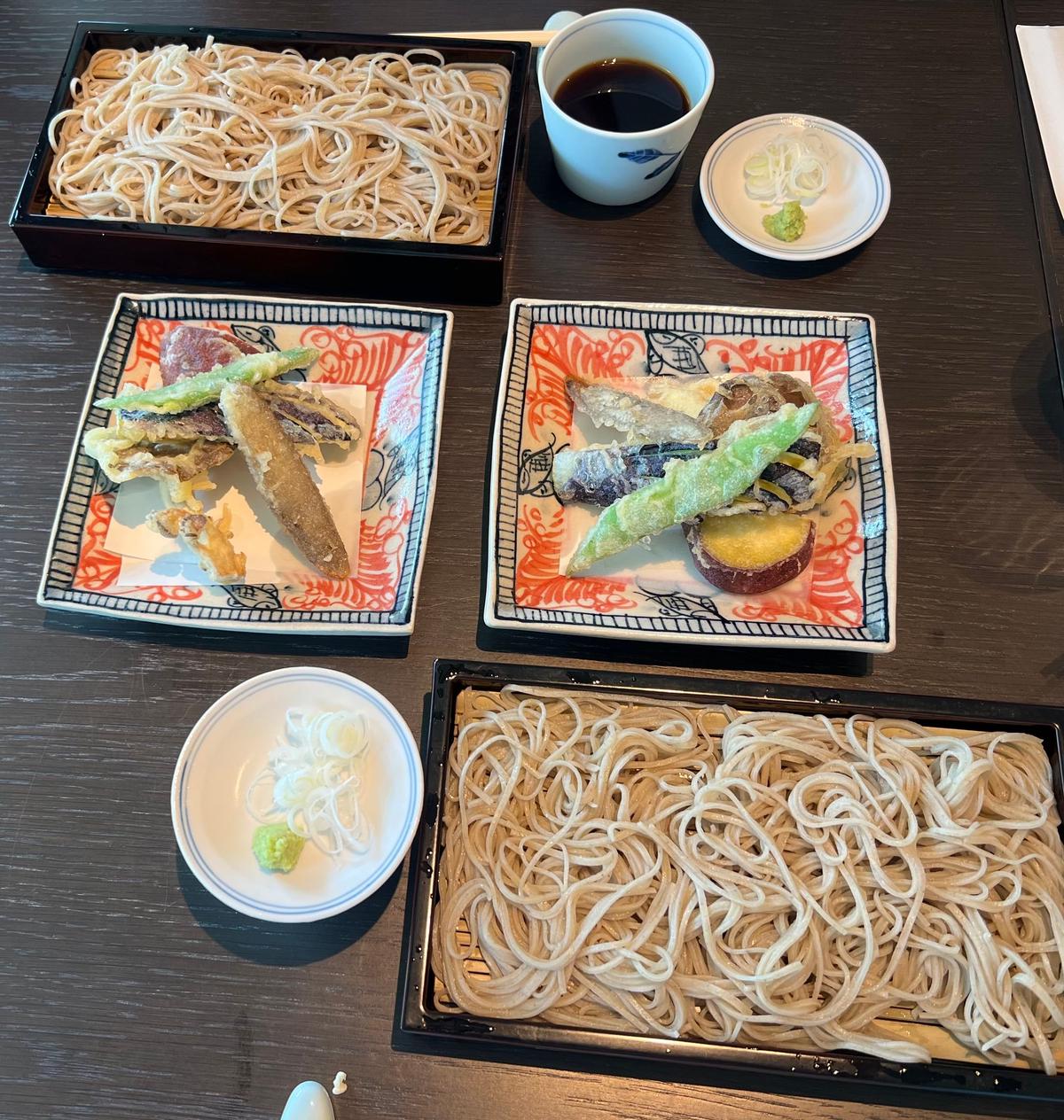 木製テーブルに、うどん2皿、揚げ物2皿、豆腐の盛り合わせ、わさびとごまの小皿、コーヒーカップが並んでいる