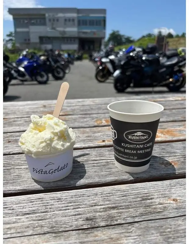 木製のテーブルに置かれた、ピスタチオジェラートと紙コップに入ったコーヒー。背景にはバイクが並び、晴天の空が広がっている。紙コップには「KUSHITANI CAFE COFFEE BREAK MEETING」のロゴ。