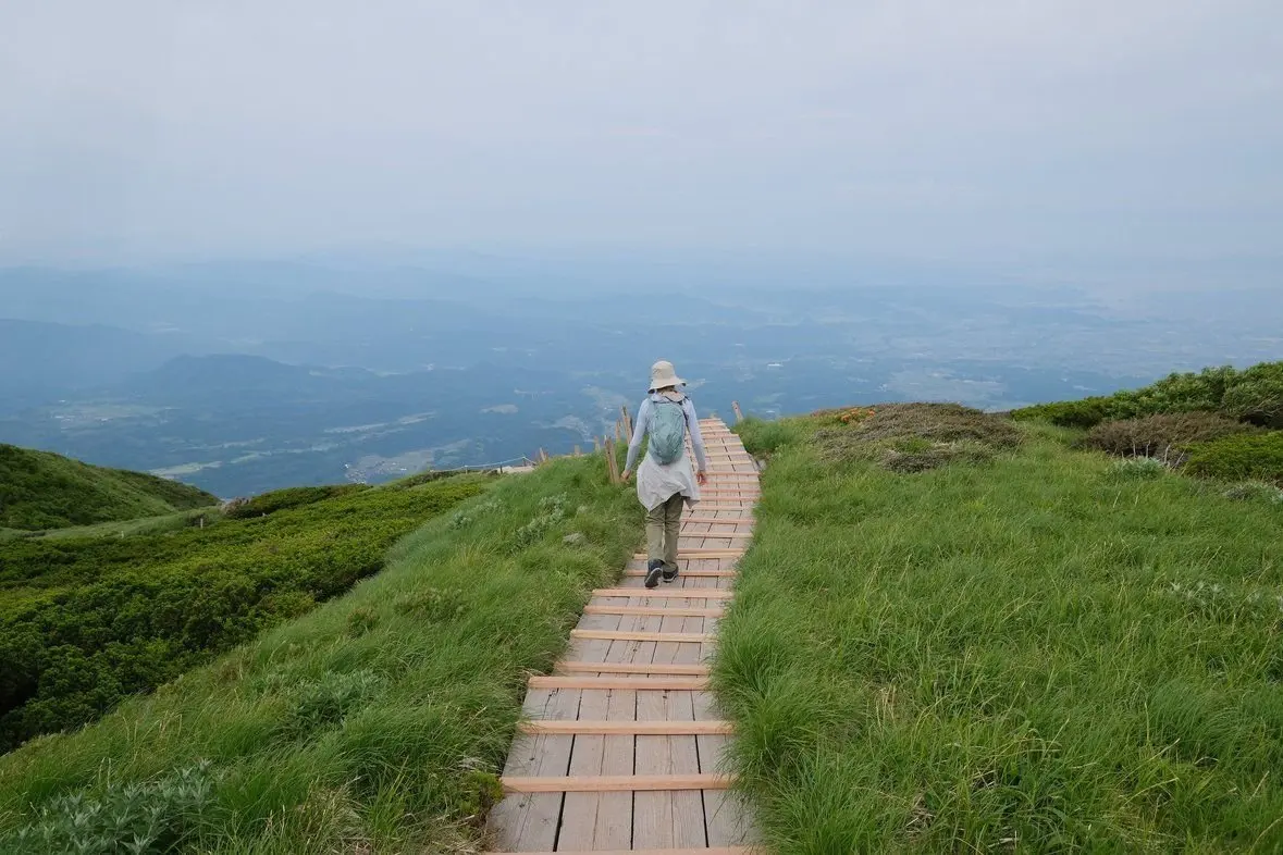 山頂の木製階段を歩く女性、広大な山並みと遠くの街並みが背景に広がる