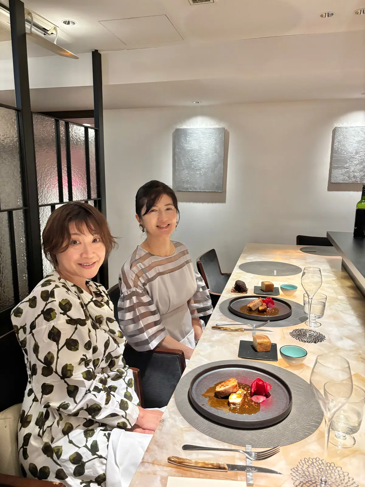 二人の女性がテーブルに座り、料理とグラスが置かれたレストランのテーブルを前にして微笑んでいる。