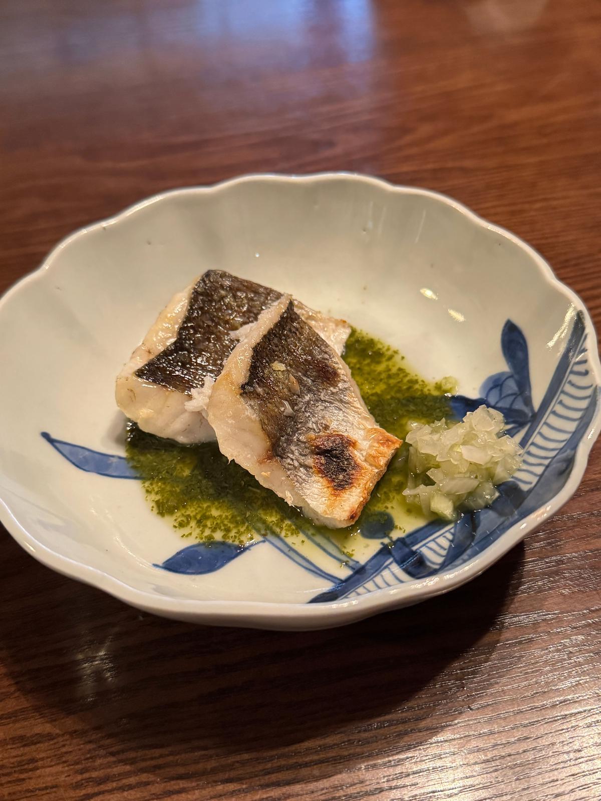 白地に青い葉模様の和風の小皿に盛り付けられた、香ばしい焼き魚。魚は白身で、表面に焼き色がついており、緑色の薬味と白っぽい和え物が添えられている。