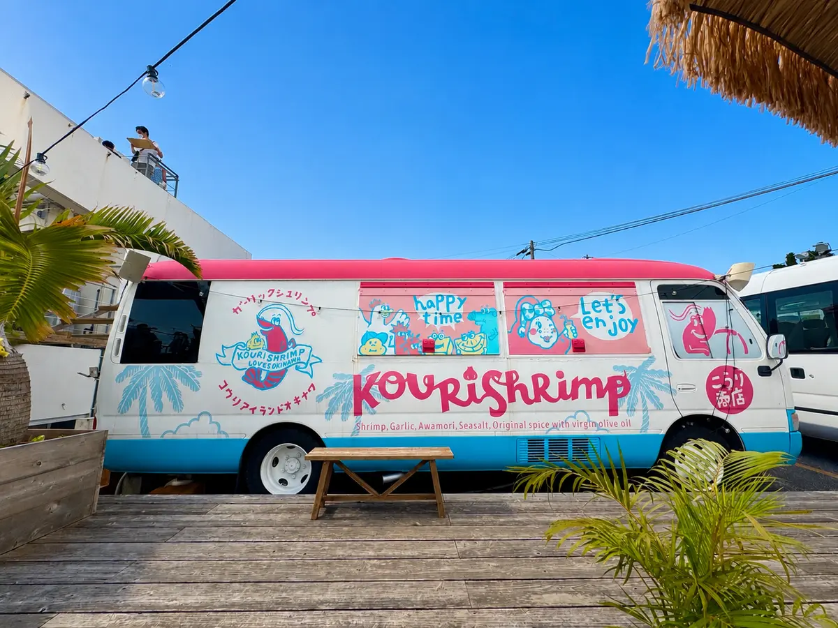エビ料理の移動販売車「Kourishrimp」がピンクと青のデザインで、木製のテラスに停車している。車体にはエビやパームのイラストと日本語の文字が描かれている。