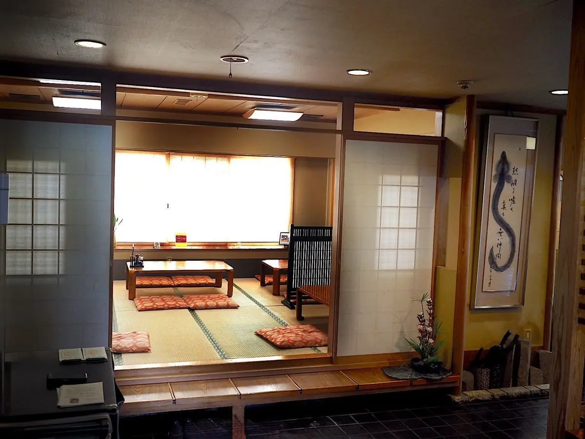伝統的な日本の部屋。障子で仕切られた空間に、畳敷きの床、ローテーブル、座布団が配置。右壁には書と植物の飾りが、左側には格子戸がある。