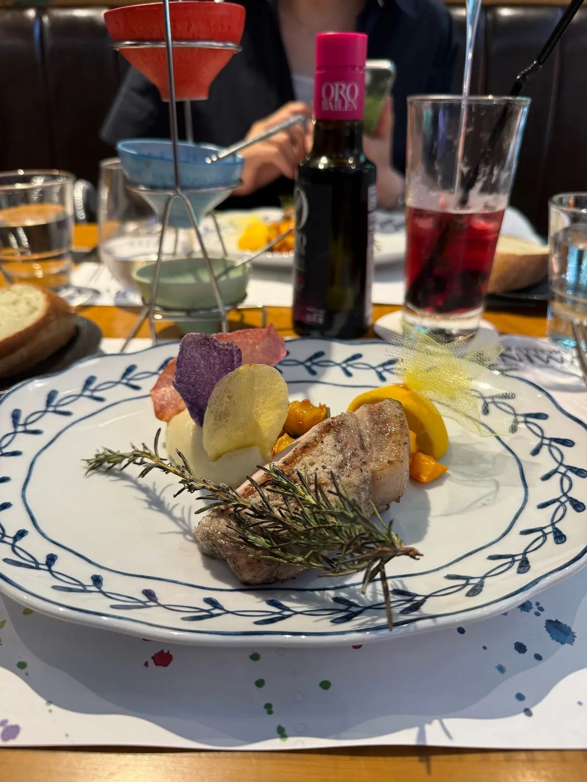 表参道レストラン、イタリアンレストラン庭、白い器に、ローストした魚の切り身、ポテトのスライス、野菜の盛り合わせ、ローザマリーの花が添えられた料理。背景には赤ワインとグラスが置かれている。