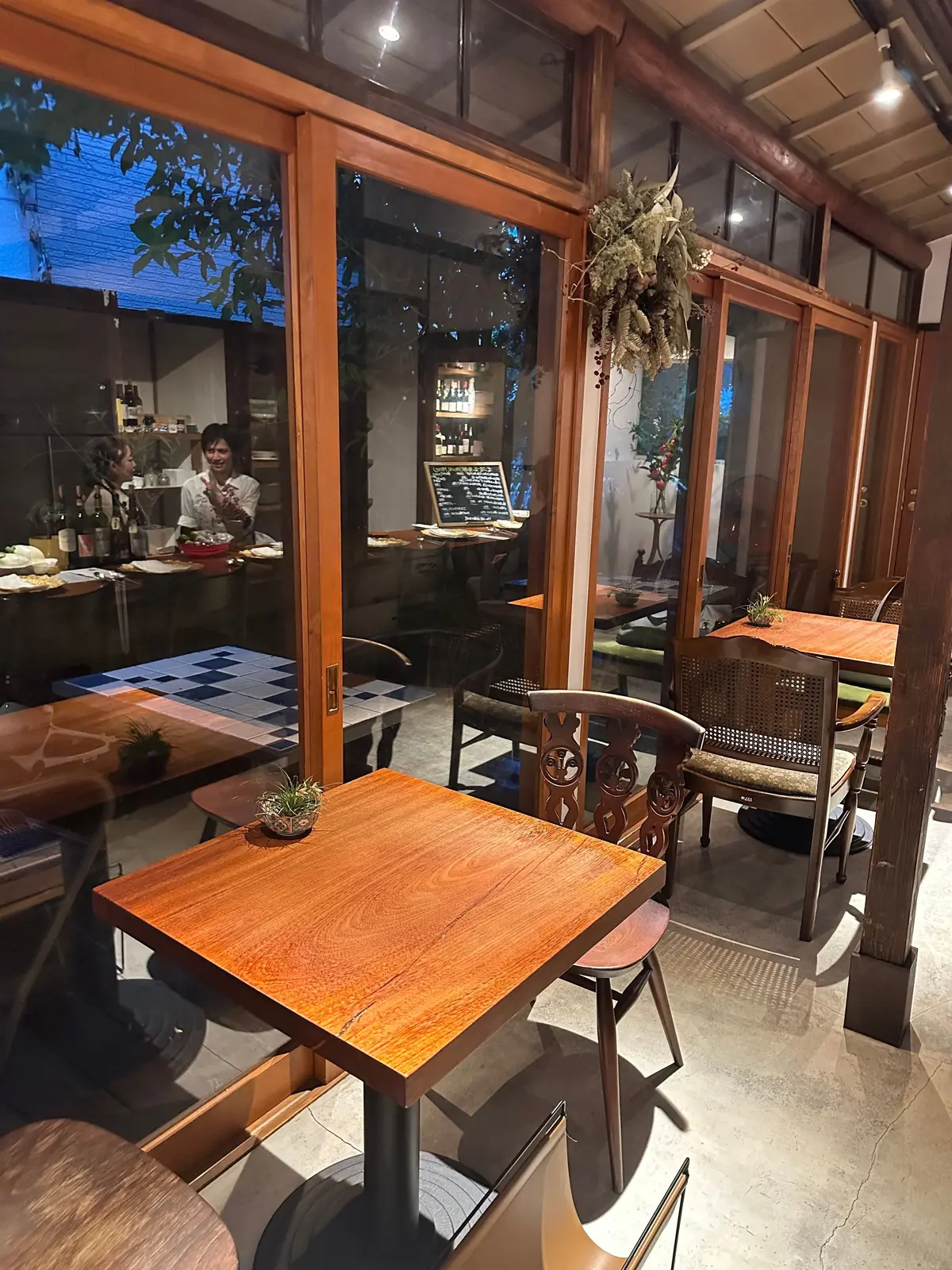 木製のテーブルと椅子が並ぶ、落ち着いた雰囲気のレストランの内部。窓の外には緑が見え、メニューが掲示されている。左奥には客が食事をしている様子も見える。