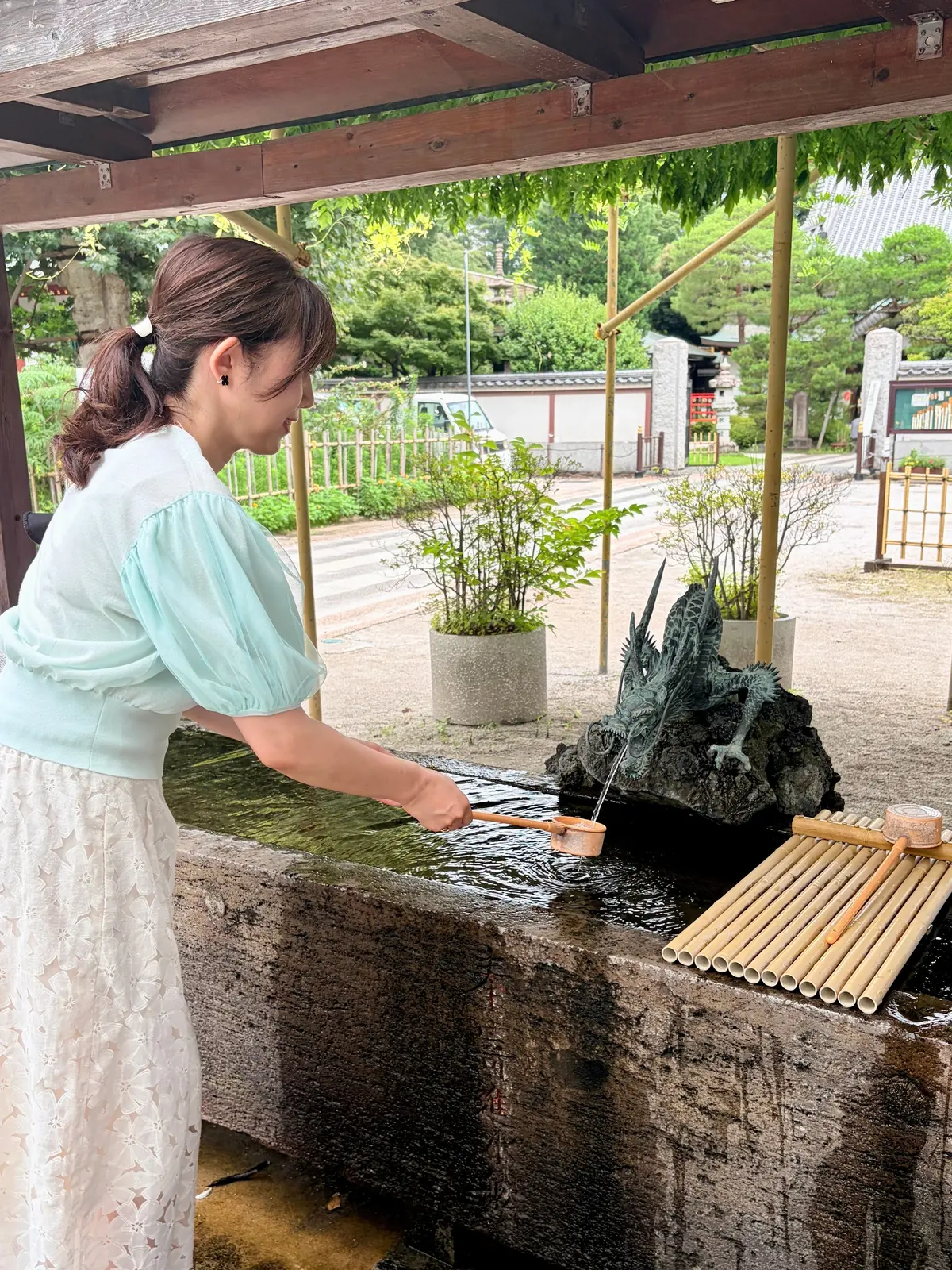 緑色のトップスと白いワンピースを着た女性が、日本の寺にある注水盤で水をくみ上げている。注水盤には龍の彫刻があり、竹製の樋と受け皿が設置されている。背景には緑豊かな植栽と建物が見える。