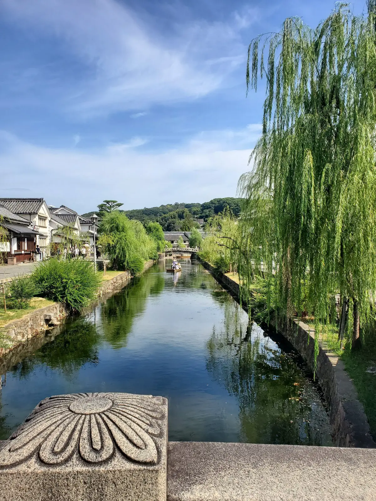 日本の風景。水路が左右に伸び、柳の木が並ぶ。背景には伝統的な家並みと緑豊かな山が見える。青空に白い雲が浮かんでいる。