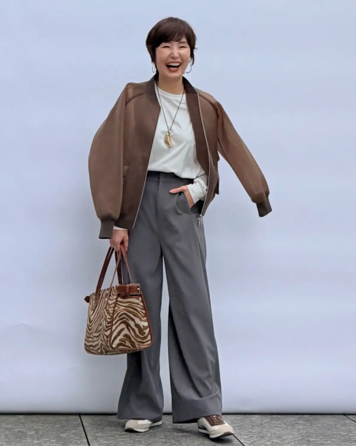 茶色のボマージャケットに白いカットソー、グレーのワイドパンツを着用した女性が笑顔で立っている全身写真。右手に茶色のゼブラ柄バッグを持ち、ネックレスを着用。背景は白い壁。