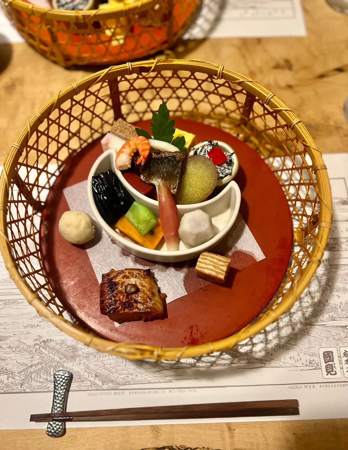 黄金色の竹製バスケットに、赤いプレートに盛られた多彩な料理。白い椀にエビ、魚、野菜、豆腐が入り、周囲には焼き魚、豆腐、野菜、串焼きなど。