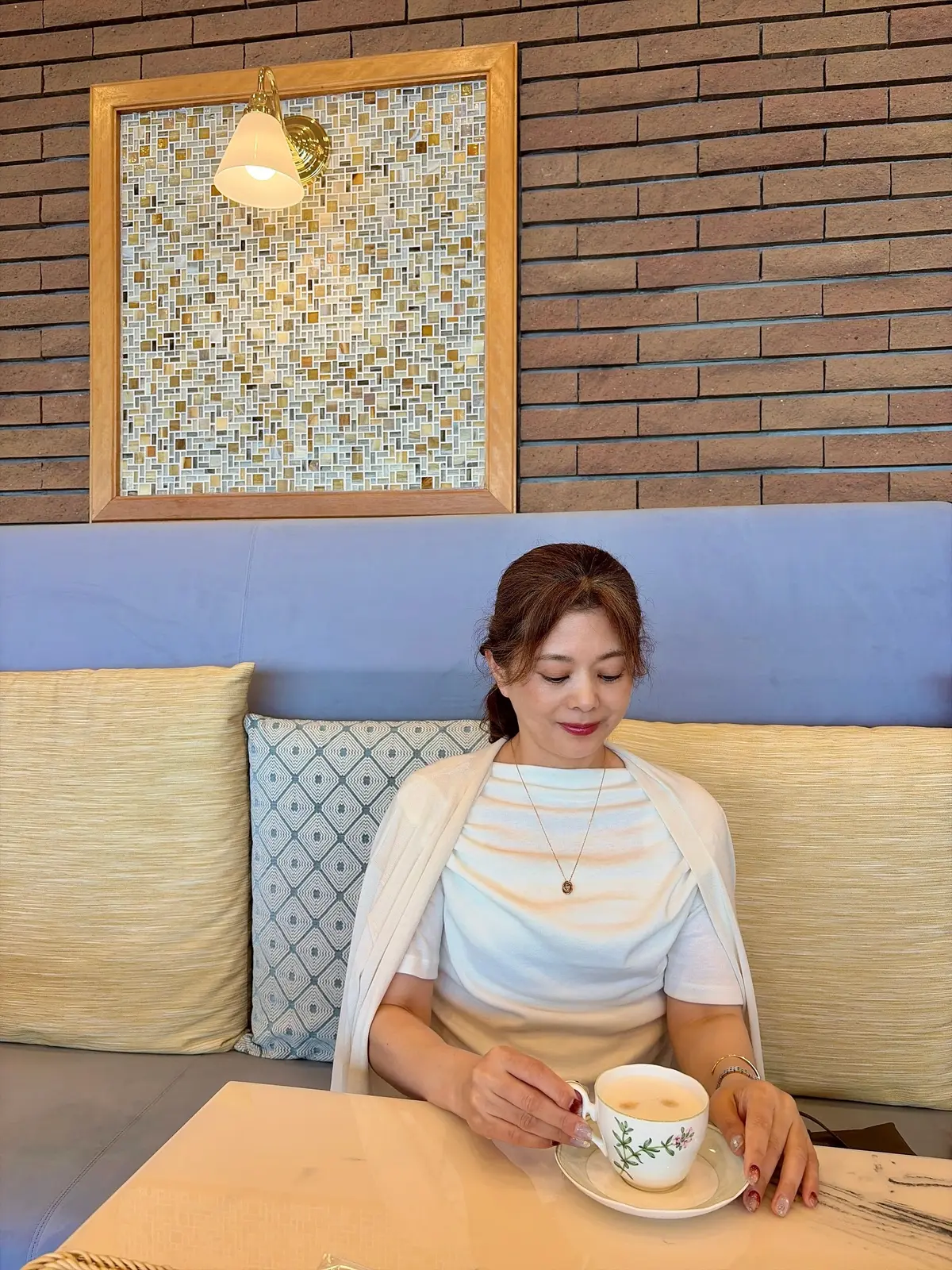 カフェのベンチに座り、白いトップスに白いショールを羽織った女性が、花柄のカップに入った飲み物を手にしている。背景には、モザイクタイルとレンガの壁、青色のベンチがある。