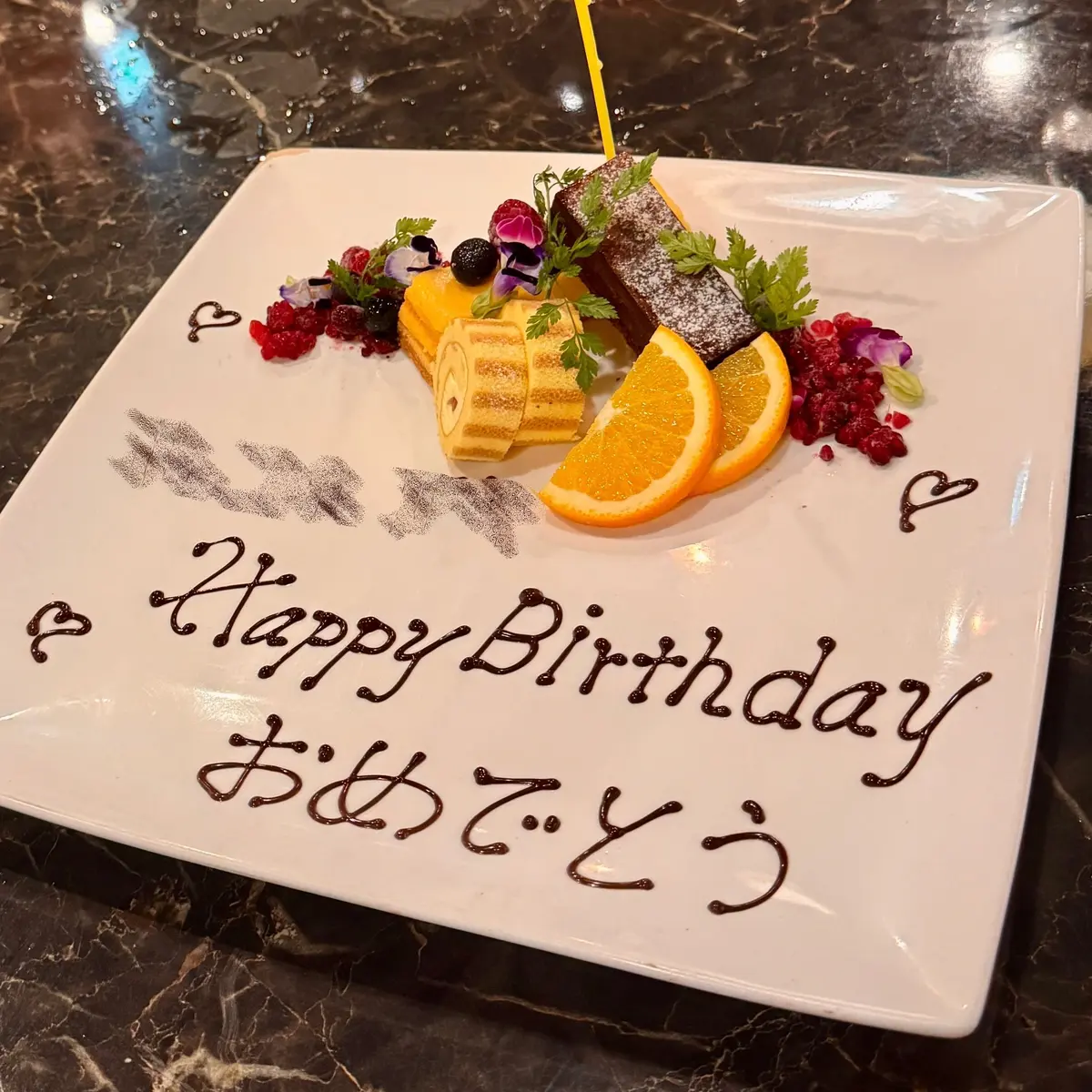 白磁の皿に、チョコレートケーキ、オレンジのスライス、ベリー、花々が盛り付けられた誕生日デザート。チョコレートケーキは縦に3切れ並び、オレンジは半分にカットされ、ベリーは赤黒い色合い。