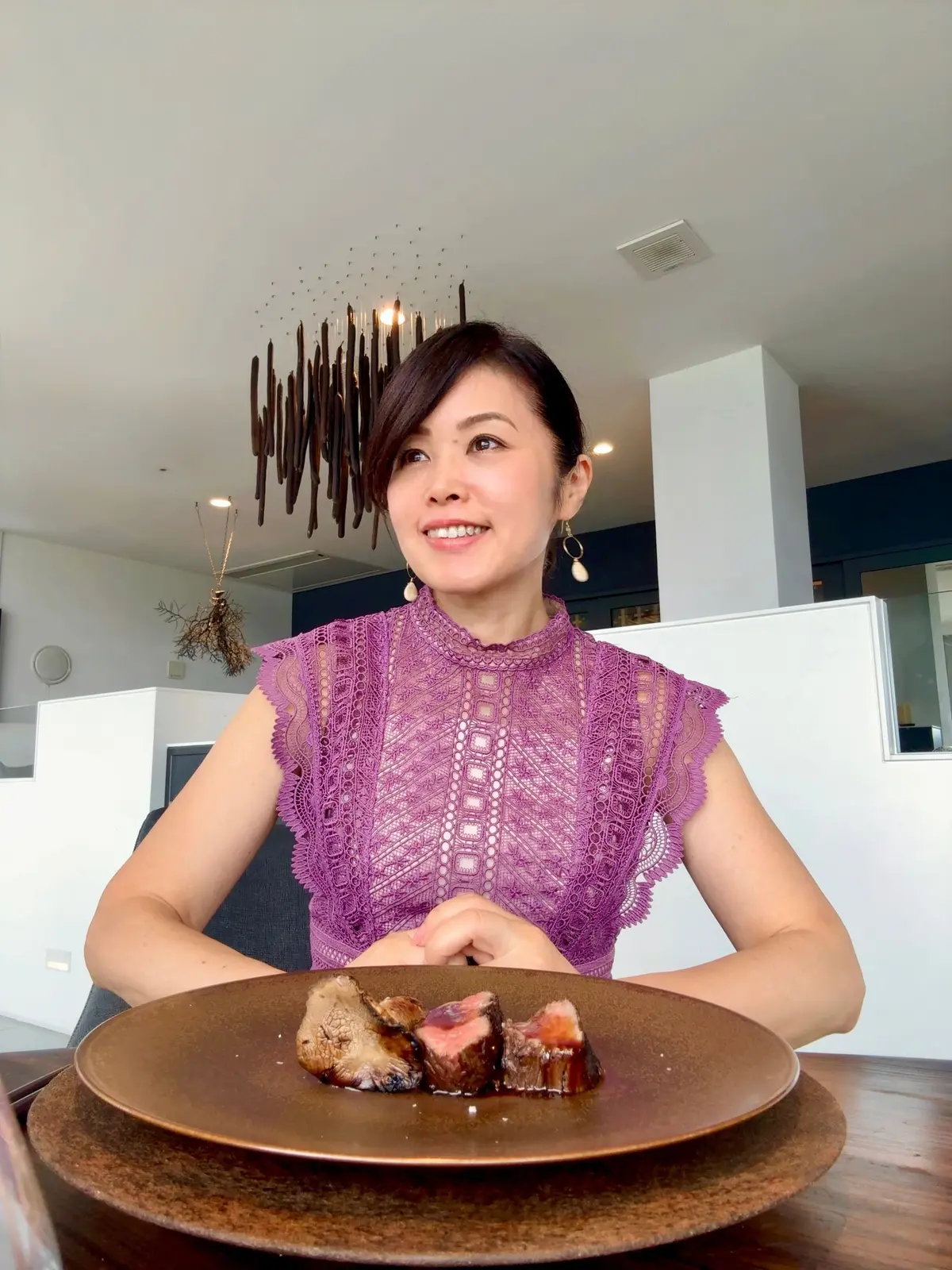 マグピンクのレース風トップスを着た女性が、木製のテーブルに置かれた焼き肉の盛り合わせを前に微笑んでいる。背景にはモダンな照明が設置されている。
