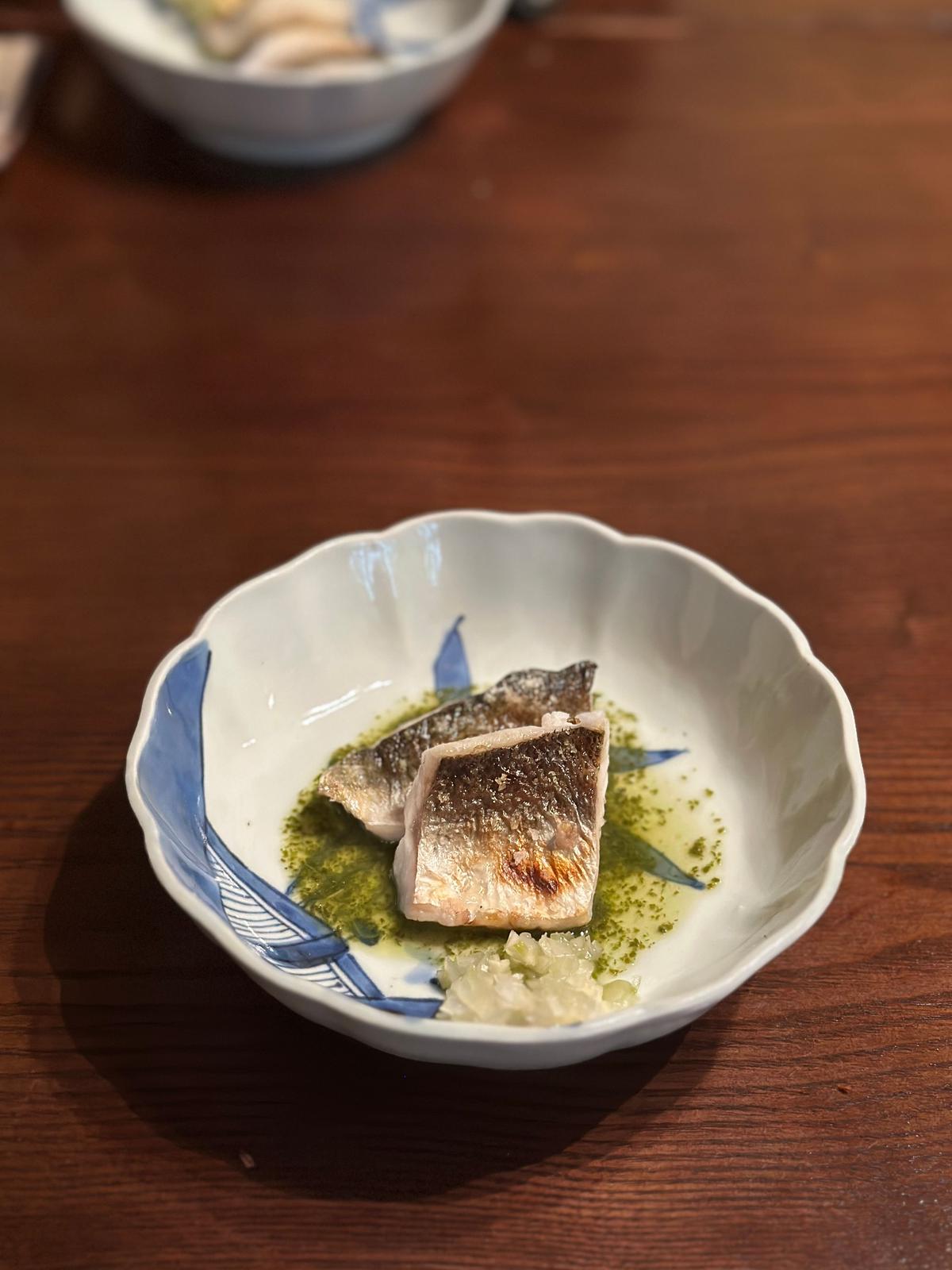 白と青の模様の小皿に盛り付けられた、照り焼き風味の魚の切り身。魚の表面は焼き色が付いており、皿には緑色のソースとすりおろしニンニクが添えられている。