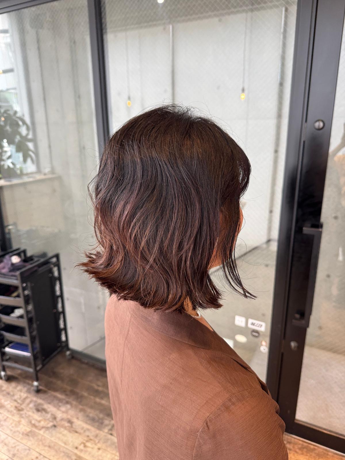 肩くらいの長さのダークブラウンのボブヘア。ゆるやかなウェーブがかかり、光沢がある。背景には、美容室のドアと内装が見える。