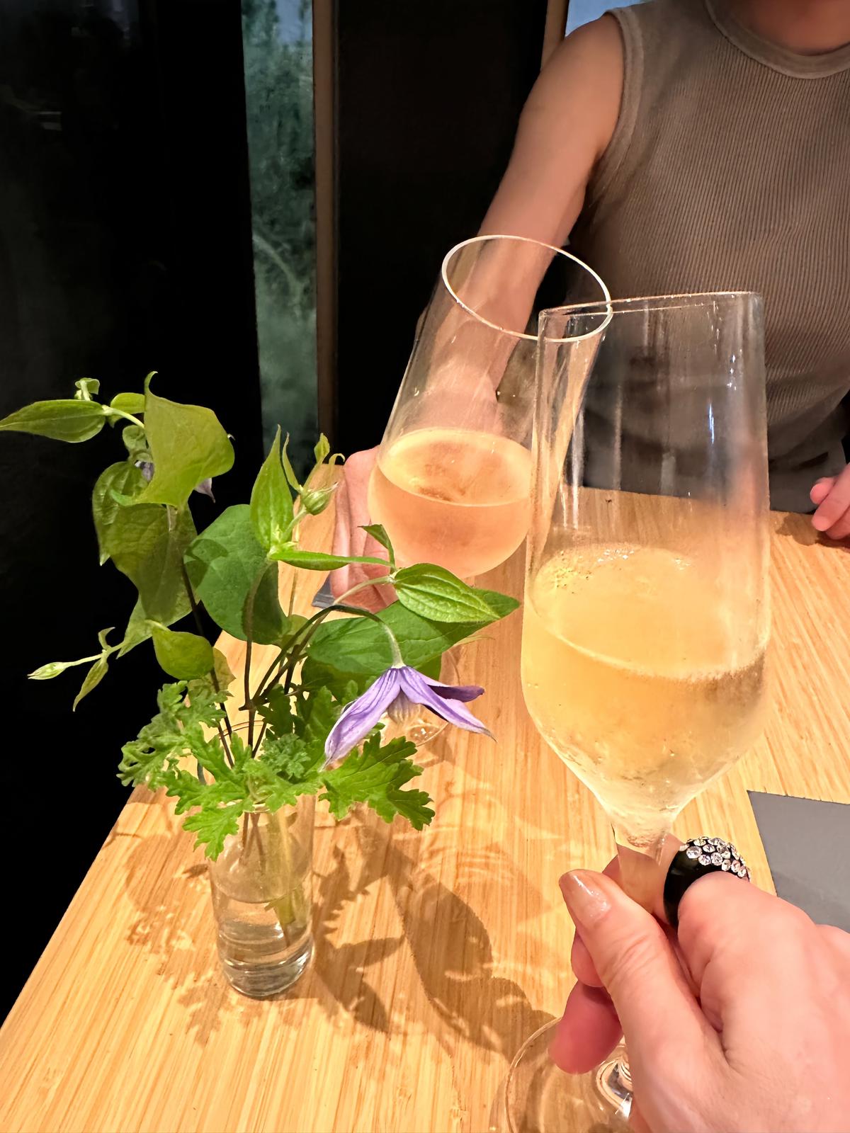木製のテーブルに置かれた、傾いたグラス2つに入った淡いオレンジ色のスパークリングワイン。小さな花瓶には緑の葉と紫色の花が飾られている。手はグラスを握り、背景には人物と暗い壁が見える。