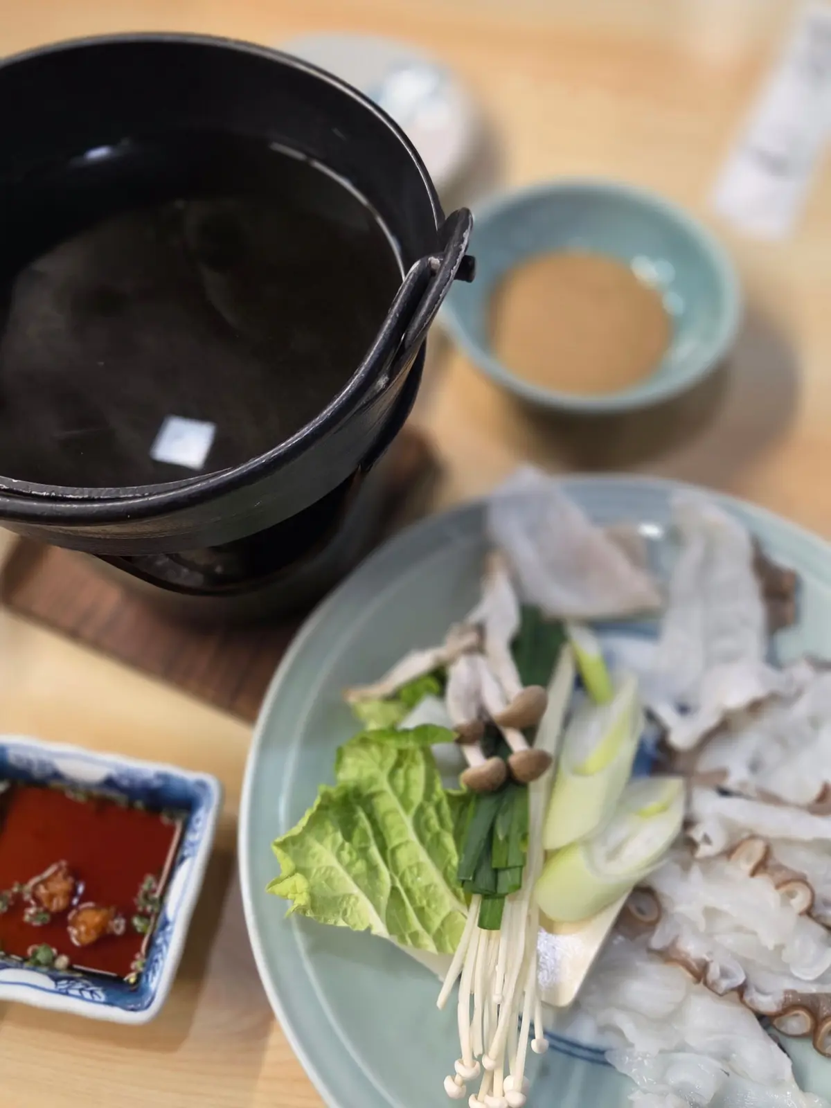 木製のテーブルに置かれた、韓国料理のセット。黒い器にはスープ、小皿にはタレ、青い皿には生野菜、肉、茹でた麺類が盛り付けられている。