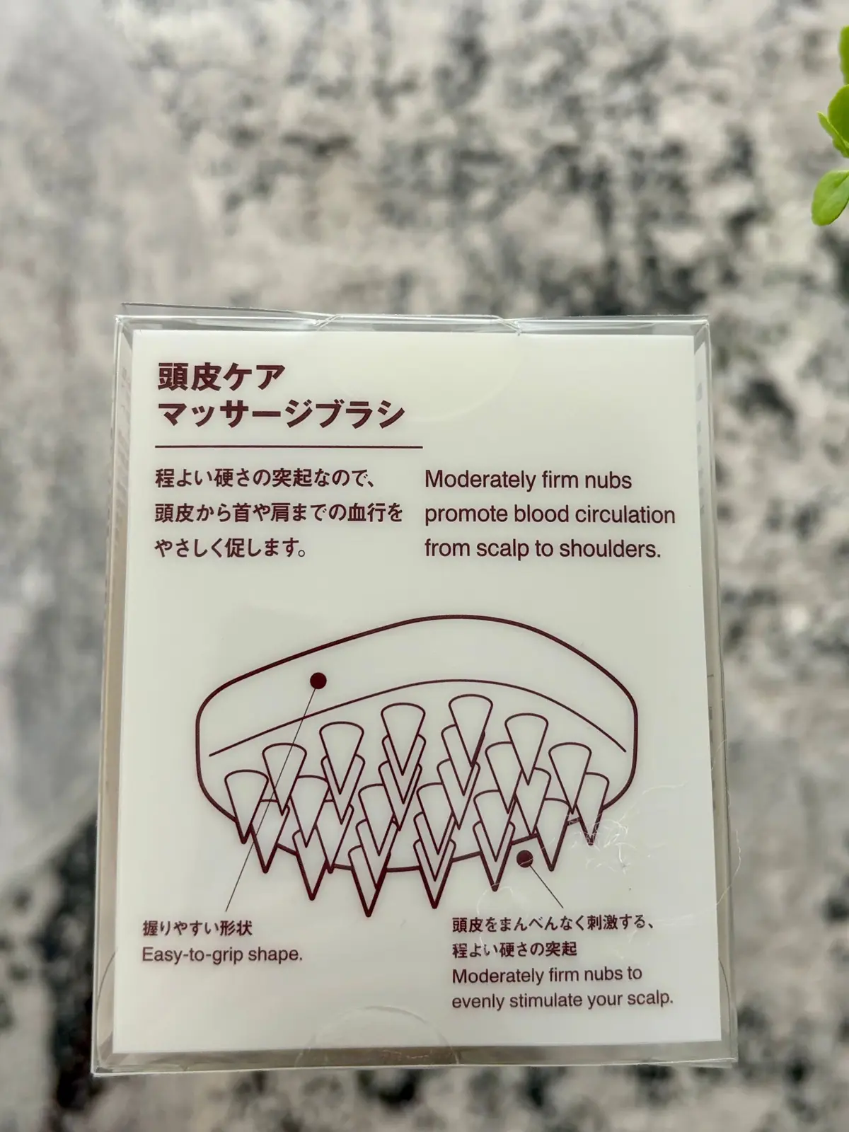 頭皮マッサージブラシ。グレーのプラスチック製で、表面に多数の丸い突起がある。製品のパッケージに、日本語と英語の説明文、イラストが記載されている。