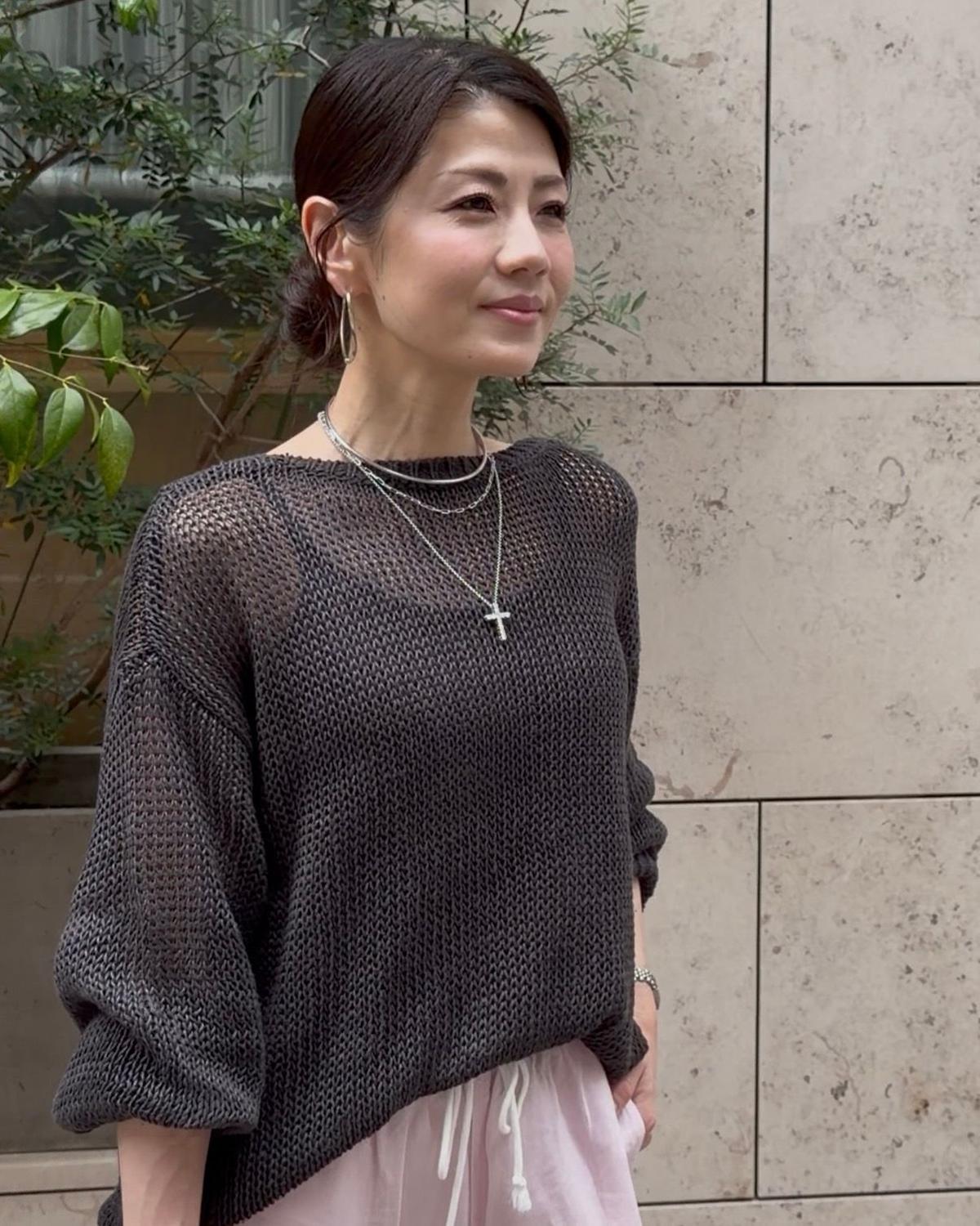 女性が左を向いて微笑んでいる。グレーの編み物セーターとピンクのスカートを着て、首には銀のチェーンと十字架のペンダントが付いている。背景には緑の植物とコンクリートの壁がある。