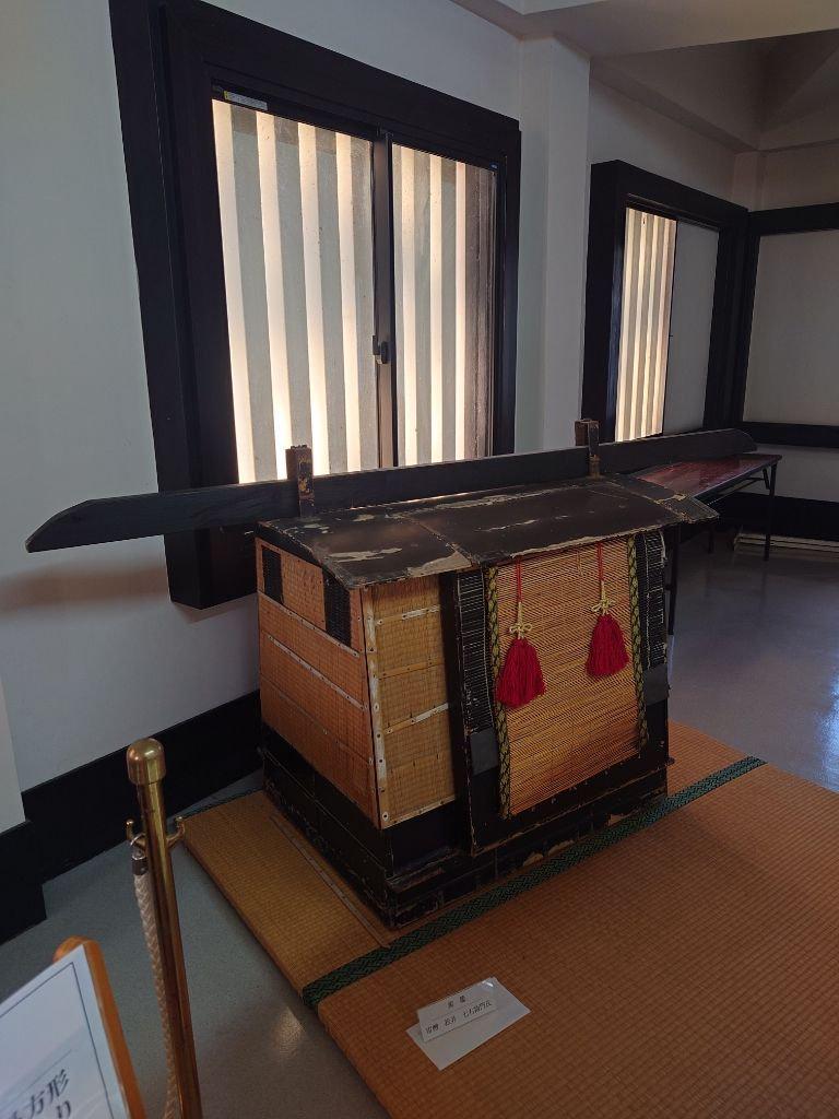 木製の箱が展示ケースに置かれている。表面は藁のような素材で覆われ、赤い房飾りが付いている。背景には、縦長の窓が左右に並んでいる。