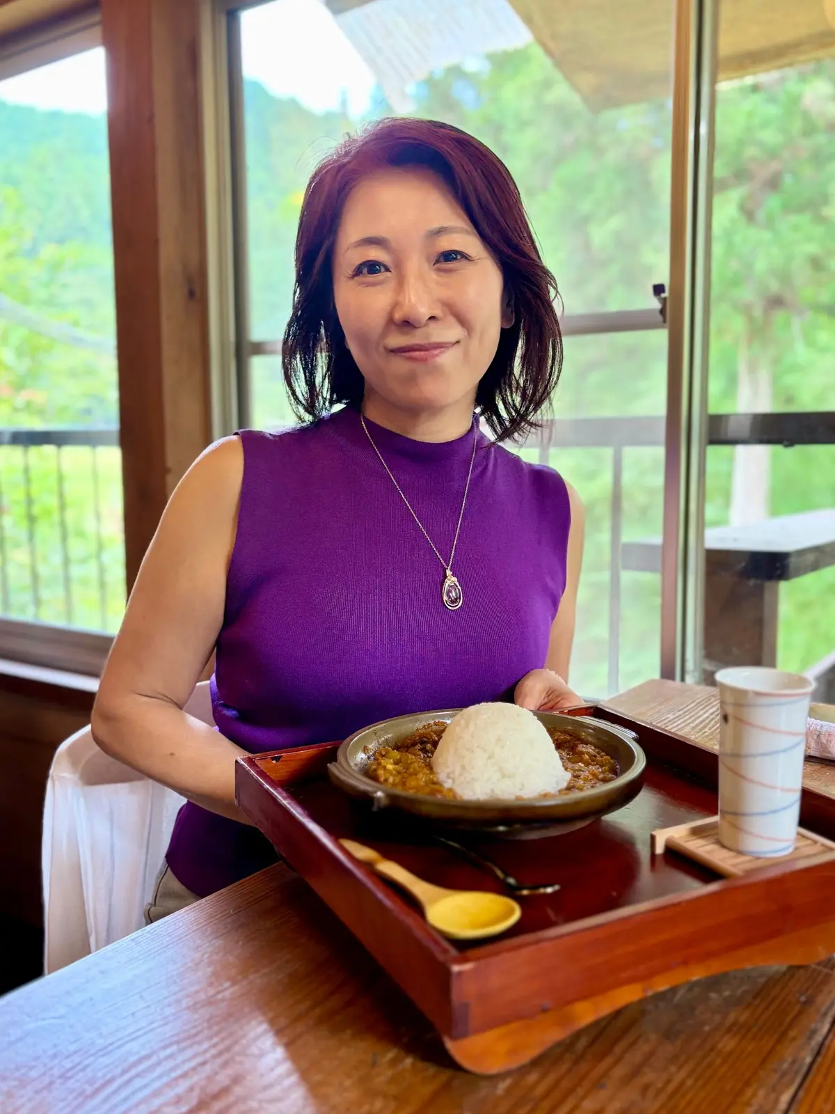 紫色のタンクトップを着た女性が、木製のトレイに乗ったカレーライスを前に微笑んでいる。背景には窓から緑豊かな景色が見える。