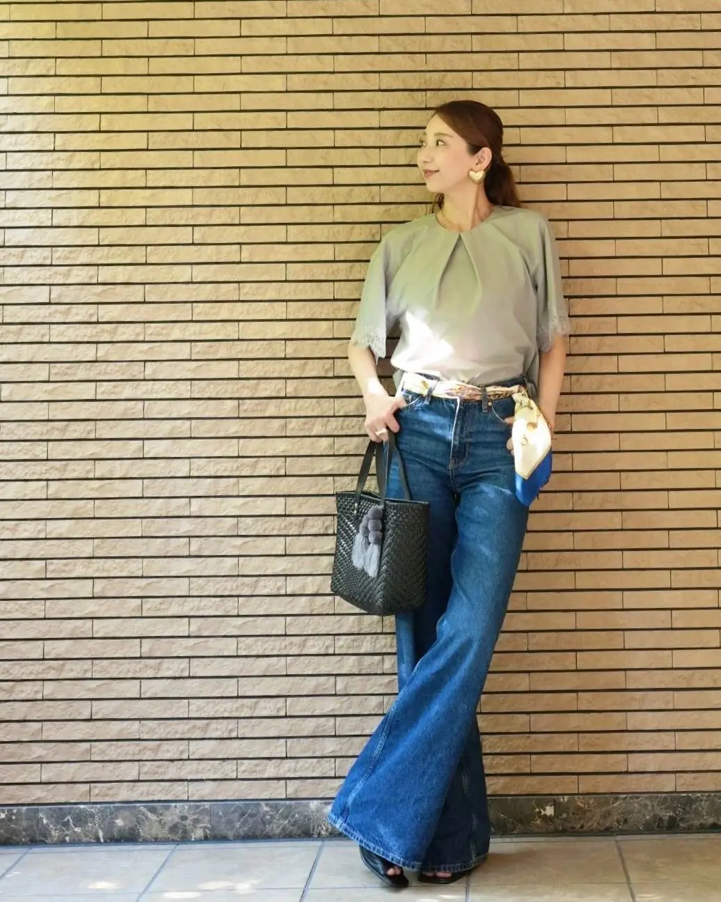 女性が横に立ち、石造りの壁に寄りかかっている。緑と白のグラデーションの半袖トップス、ジーンズ、ベルト、黒のトートバッグを手にしている。
