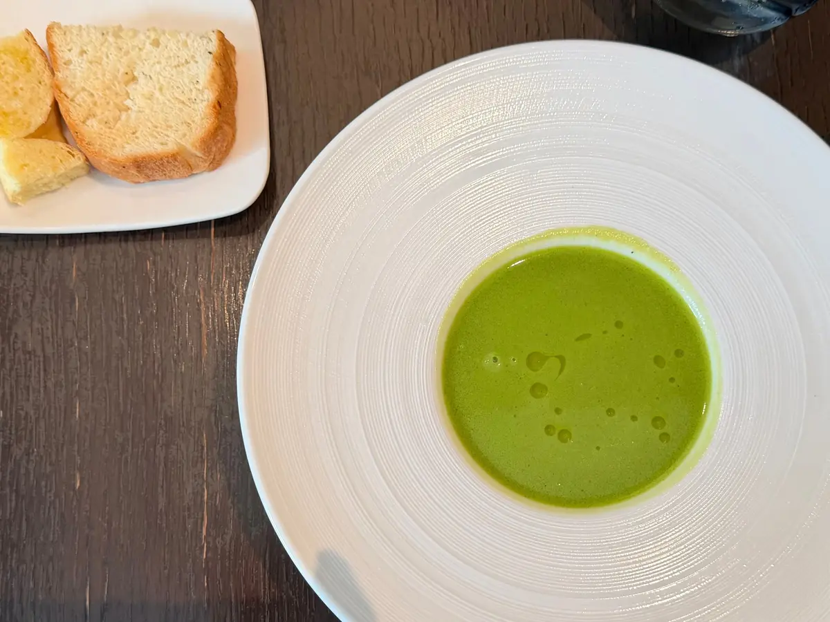 白い皿に緑色のポタージュスープ、小皿にトーストが置かれている料理の写真。木製のテーブルに配置。