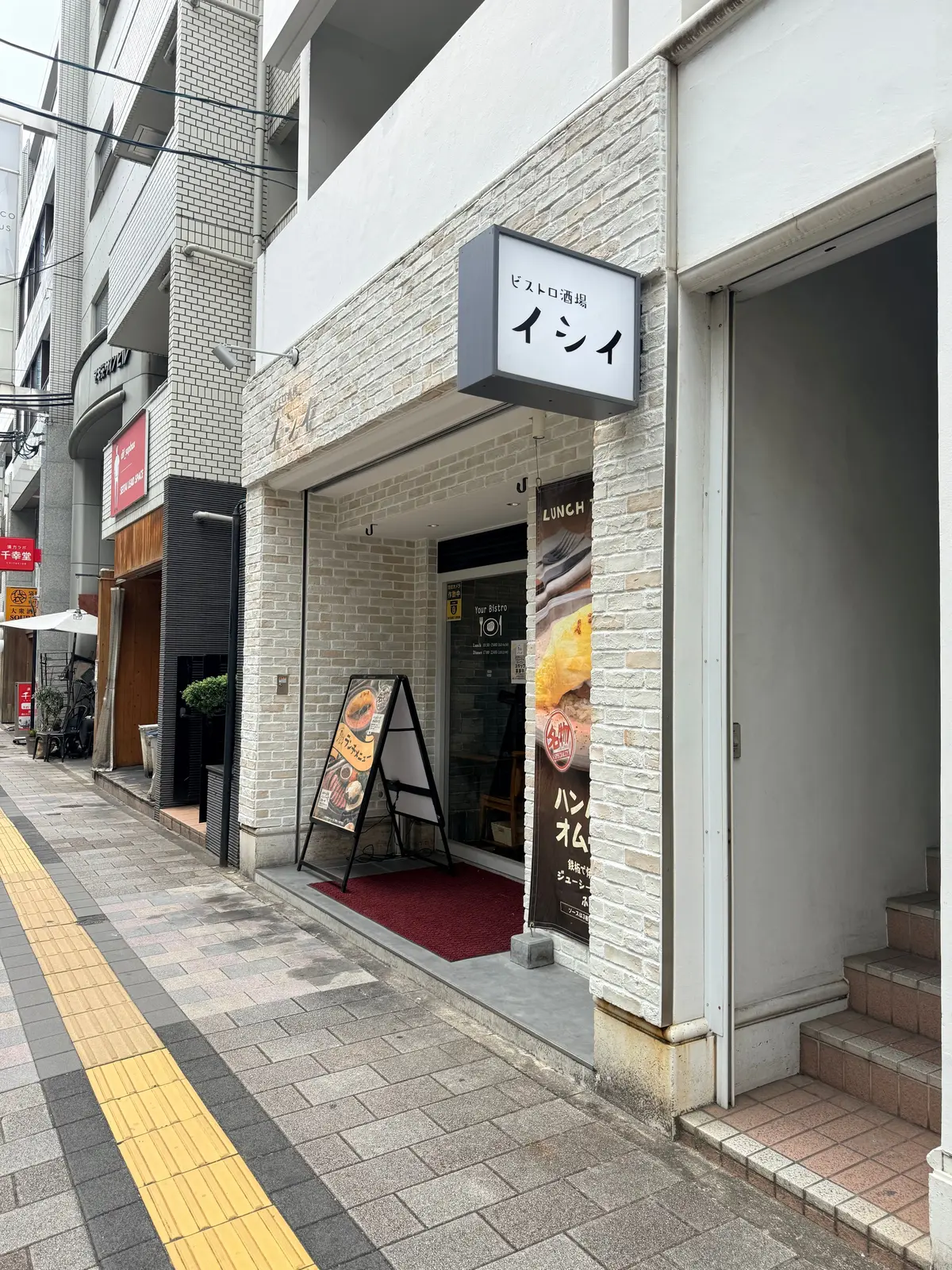 白いレンガの壁が特徴のビストロの入口。店名表示とランチメニューの看板があり、赤い敷物で飾られた入り口。通りには段差があり、左側に歩行者用黄色ラインが敷かれている。