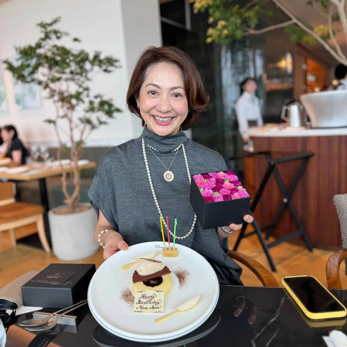 笑顔の女性が、誕生日ケーキと花束を持ってポーズをとっている。ケーキには「Happy Birthday」のメッセージが書かれている。背景にはレストランの様子が見える。