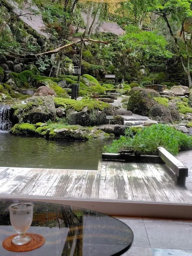moss-covered rocks and a small waterfall surround a calm pond, with a wooden pathway leading through the garden.