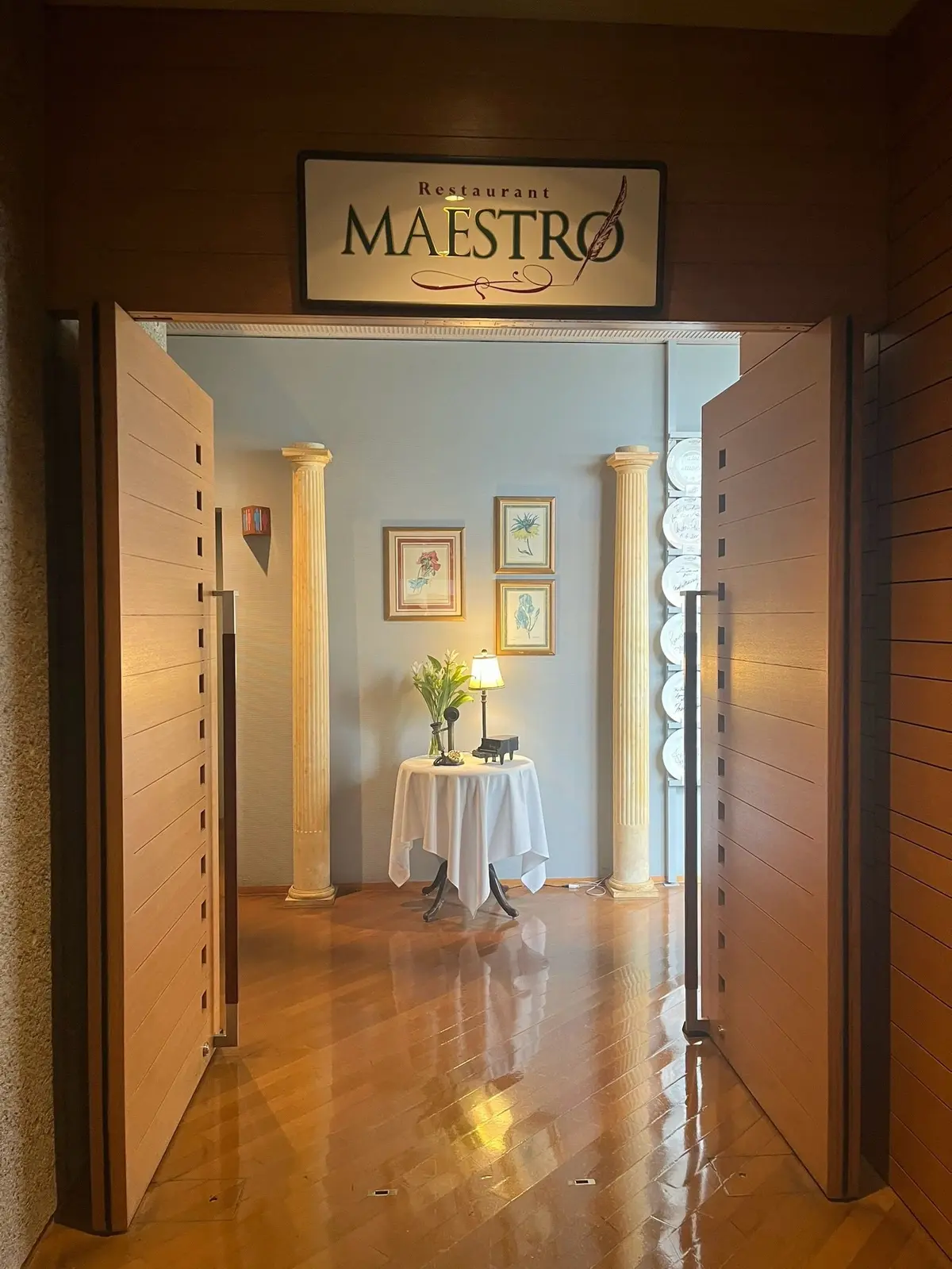 レストラン「MAESTRO」の入り口。木製の引き戸が開き、柱で仕切られた空間が見える。中央にはテーブルとランプ、植物が置かれ、壁には絵画が飾られている。床は木製で、上品な雰囲気を醸し出している。