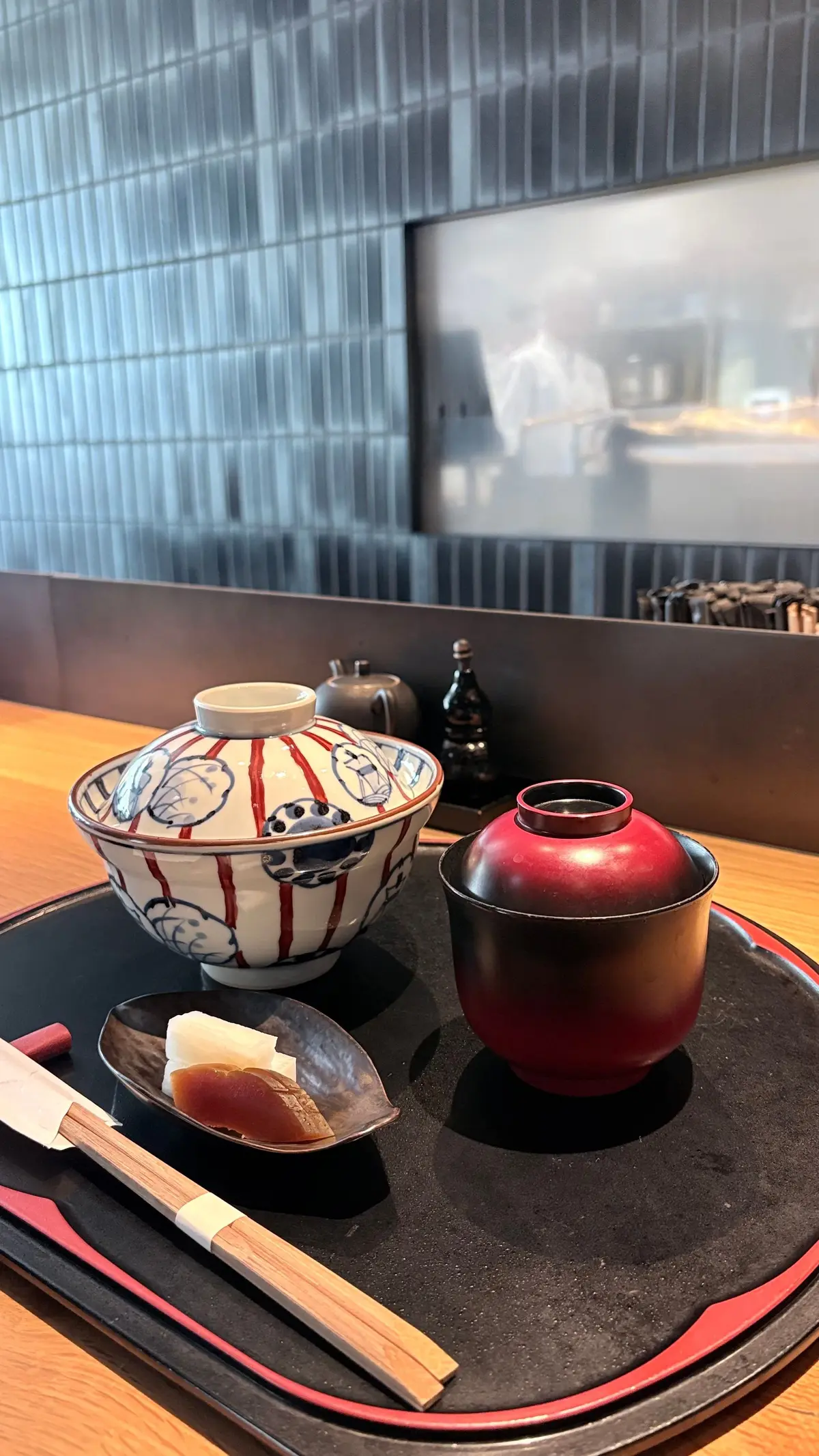 黒い漆塗りの長方形の盆の上に、白地に赤い模様の蓋付き丼、赤い汁椀、茶色の箸、そしてお新香が置かれている。背景には、青い縦縞の壁と窓の外の風景が見える。