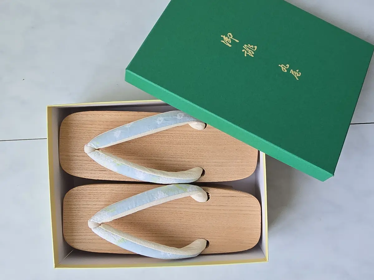 木製のゲタと、それに合わせた柄の鼻緒が特徴的な履物。緑色の箱に入っており、箱には白文字で漢字が書かれている。