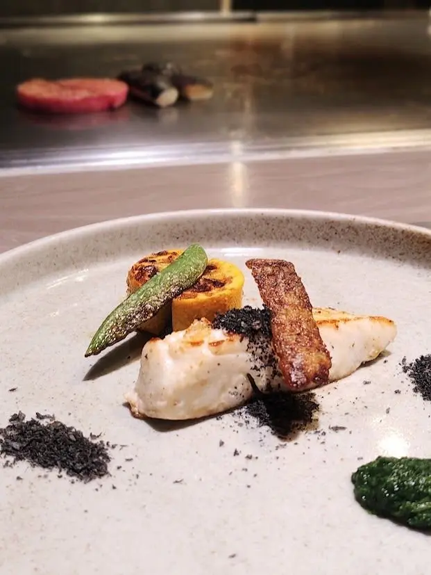 白磁の皿に盛り付けられた、白身魚のグリル料理。オレンジのスライス、緑色の野菜、焦げ目のついた棒状の食材が添えられ、黒い粉末と緑色のソースが点々と置かれている。背景には鉄板で調理された食材が見える。