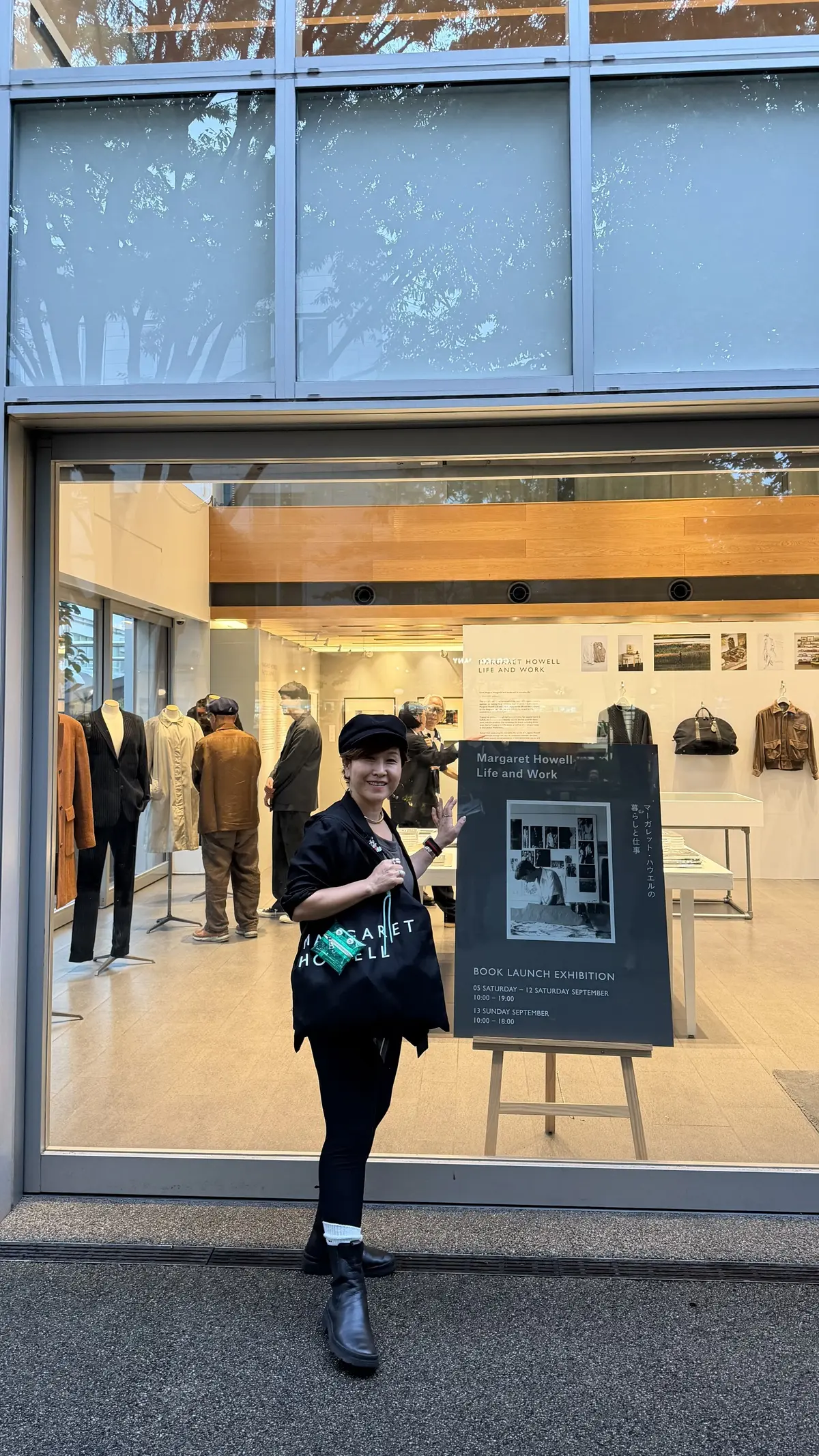 黒い帽子を被った女性が、マーガレット・ハウエルのお店の前でポーズをとっている。右手に手を上げており、胸元にマーガレット・ハウエルのロゴが入ったTシャツを着ている。背景には店内の商品や、展示会のお知らせが立てられている。