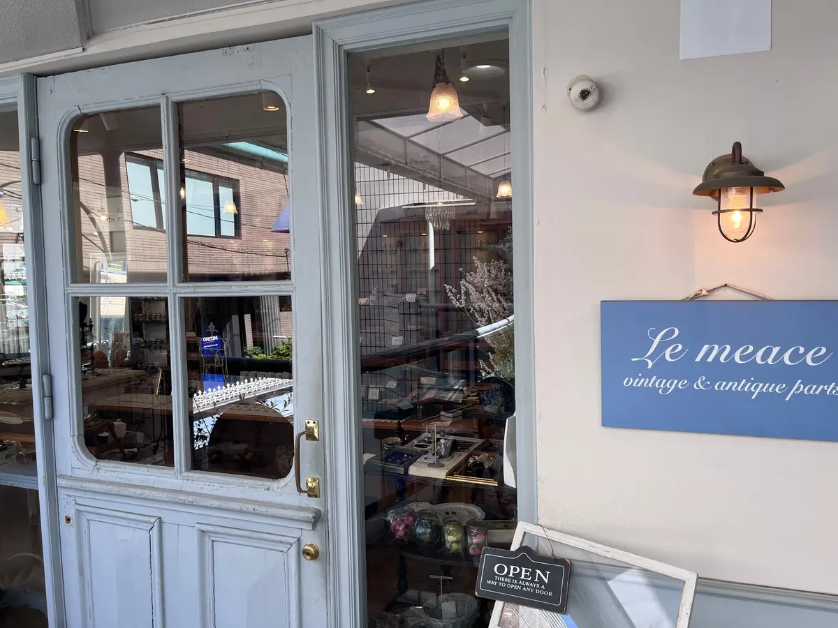 白く塗られたドアが開き、店内が見える。店内にはアンティーク家具や雑貨が並び、温かい照明が灯されている。「Le meace vintage & antique parts」の看板が右側に設置されている。