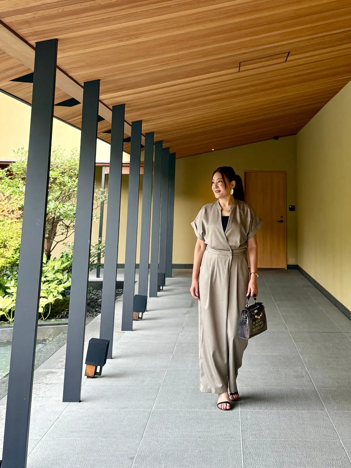 ベージュのジャンプスーツを着た女性が、灰色の通路を歩いている。木目調の天井と黒い柱が特徴的な空間。右手に黒いカバンを持ち、横にはサンダル。背景には植栽が見える。京都の大人のリゾート。ラグジュアリーホテル。