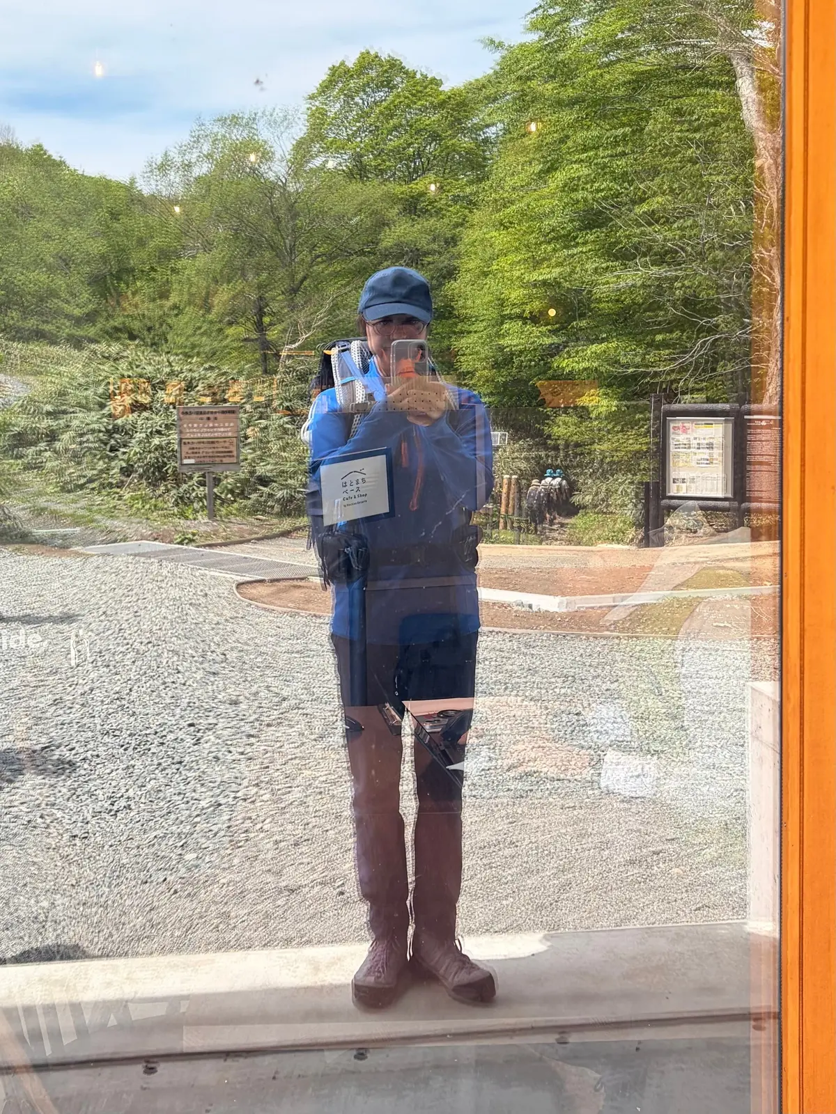 ブルーの帽子と服を着た男性が、屋外でスマートフォンでセルフポートレイトを撮っている。背景には緑豊かな木々や情報掲示板が見える。