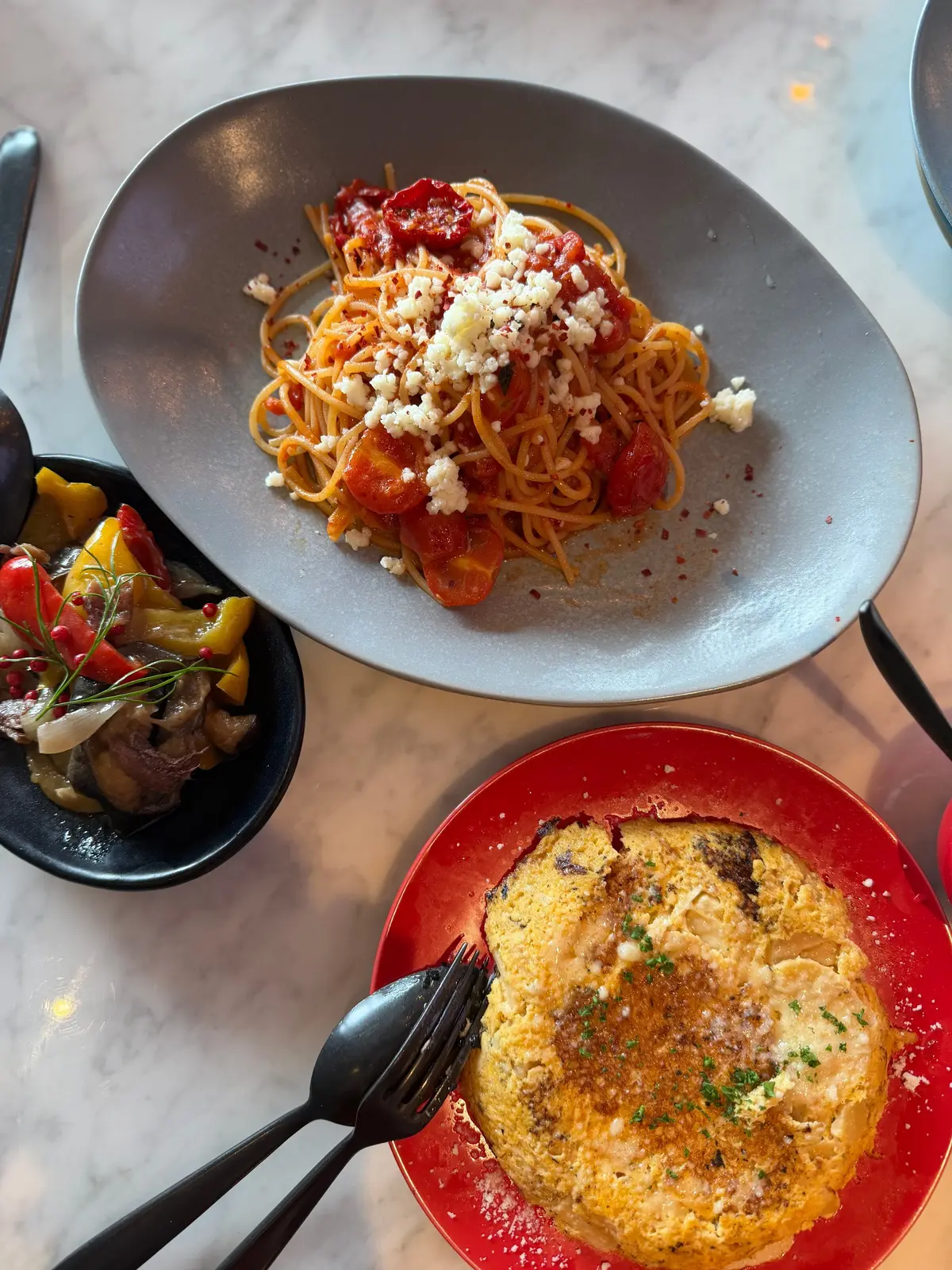 白いテーブルに置かれた3つの料理。グレーの皿にはトマトソースとチーズをかけたパスタ、赤の皿にはチーズが溶けた揚げ物、黒の皿には野菜の炒め物。黒いカトラリーが置かれている。