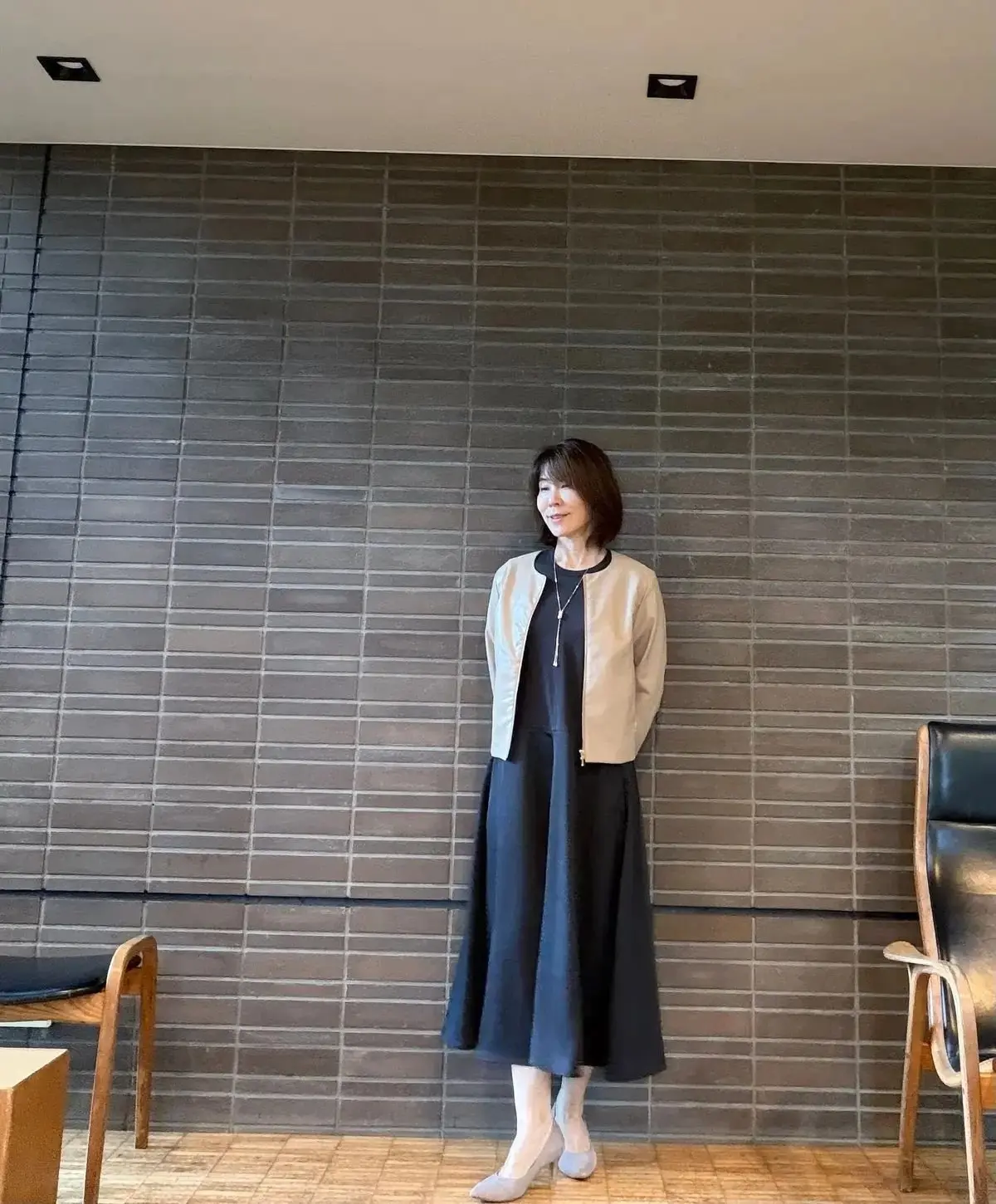 黒いミモレ丈のワンピースに、ベージュのジャケットを羽織った女性。グレーの壁を背景に立っており、灰色のパンプスを履いている。