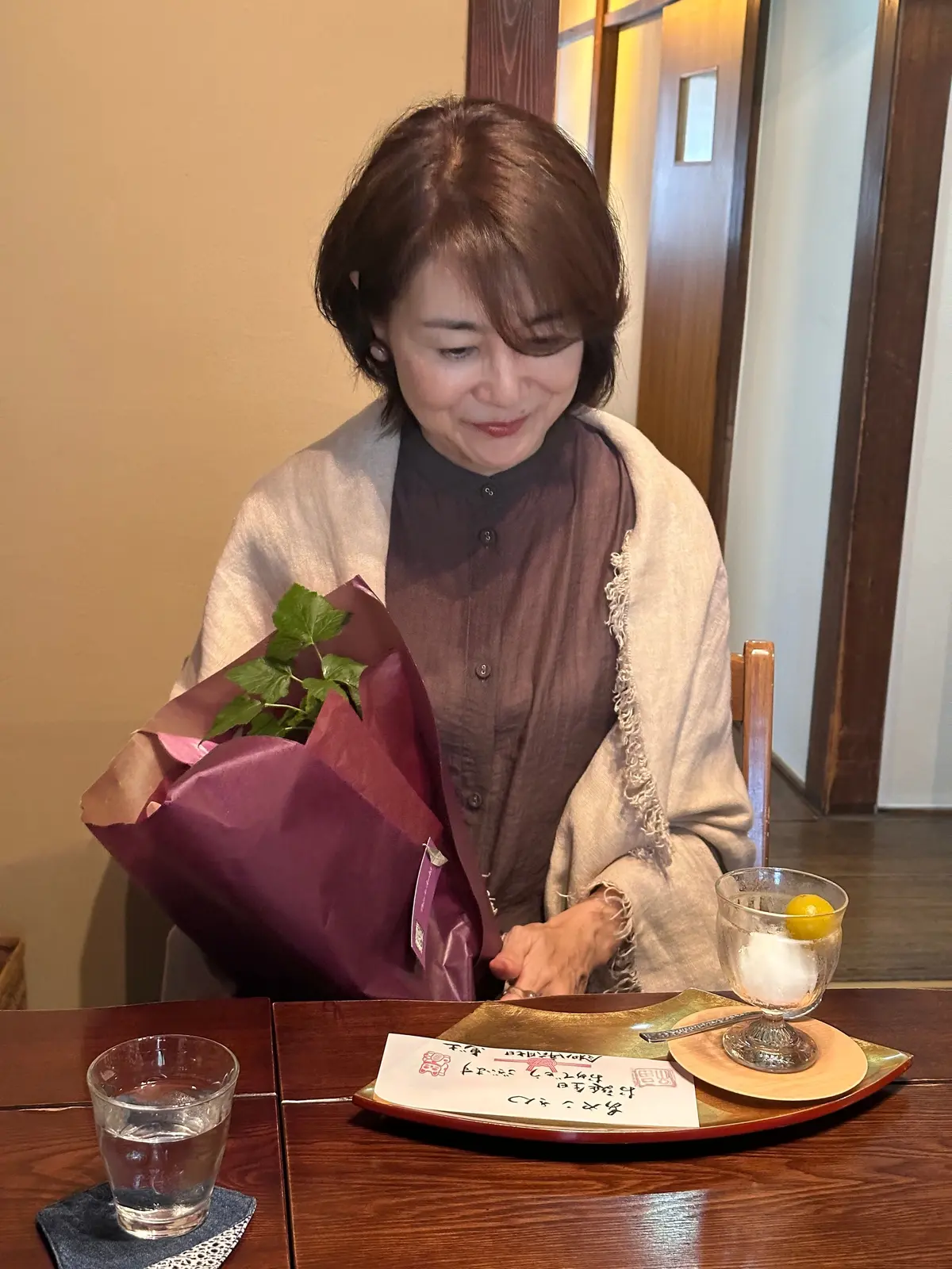 笑顔の女性が、紫色のラッピングされた花束を受け取っている様子。木目調のテーブルにはグラスと料理が置かれ、背景には木製のドアがある。