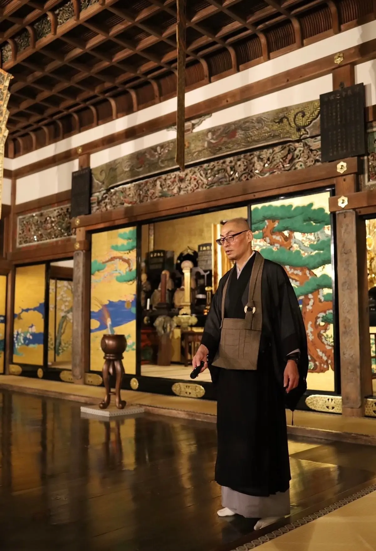 寺院内を歩く僧侶。黒い法衣を身につけ、眼鏡をかけた男性が右手を前に出し、何かを説明している様子。背景には金色の装飾画と木製の柱が見える。