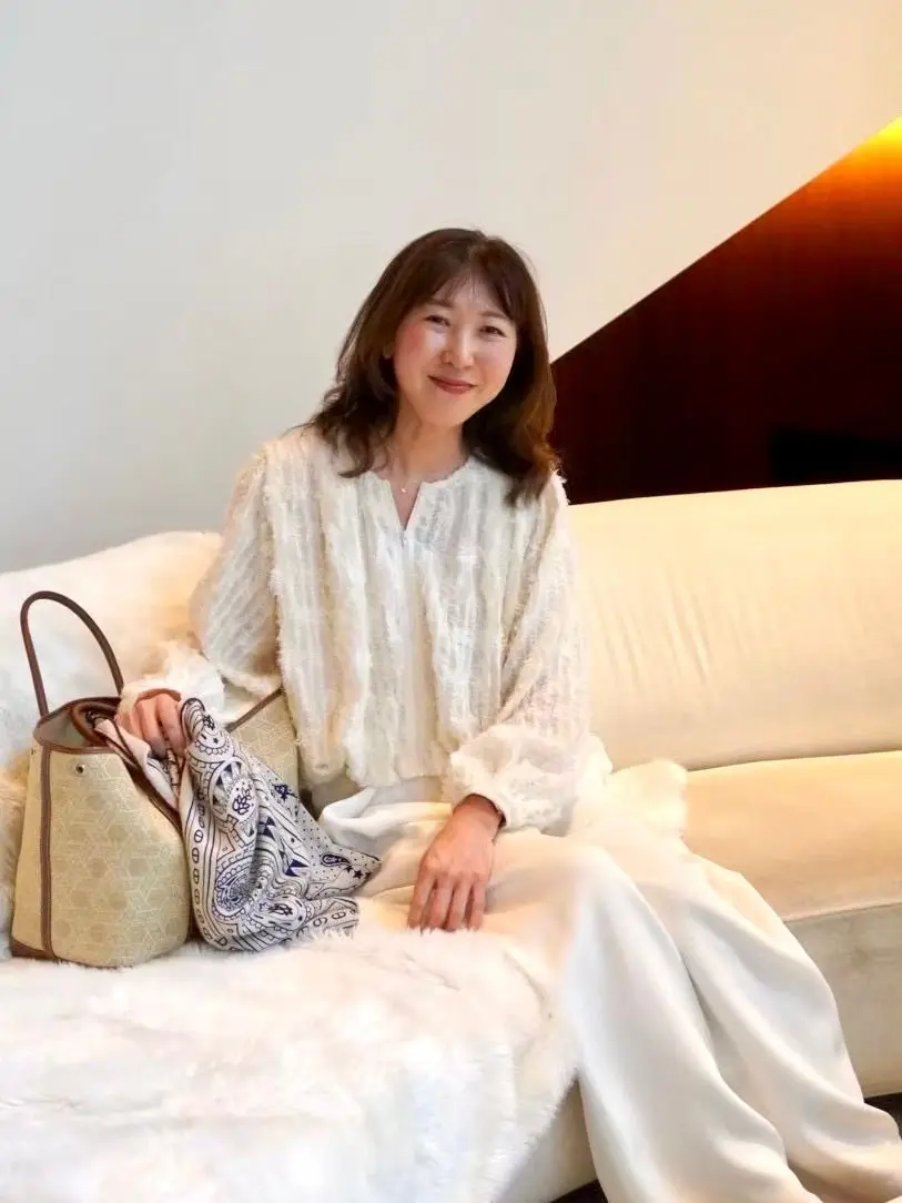 女性が白いソファに座り、笑顔で白いニットトップスを着ている。ソファの上には白いファーブランケットと、模様のあるスカーフとハンドバッグが置かれている。背景は淡い壁と木製の家具。