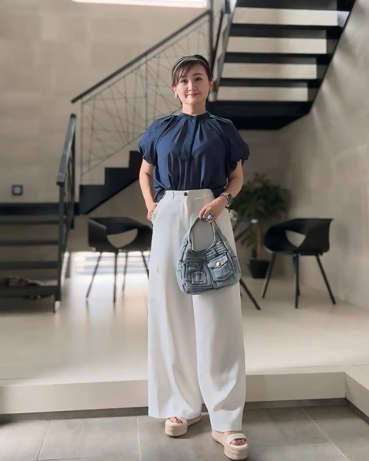 女性が室内階段の前でポーズし、紺色のプルオーバーと白いワイドパンツを着用。右手にデニム風のショルダーバッグを持ち、ベージュのフラットサンダルを履く。背景にはモダンなインテリアと植物が配置されている。
