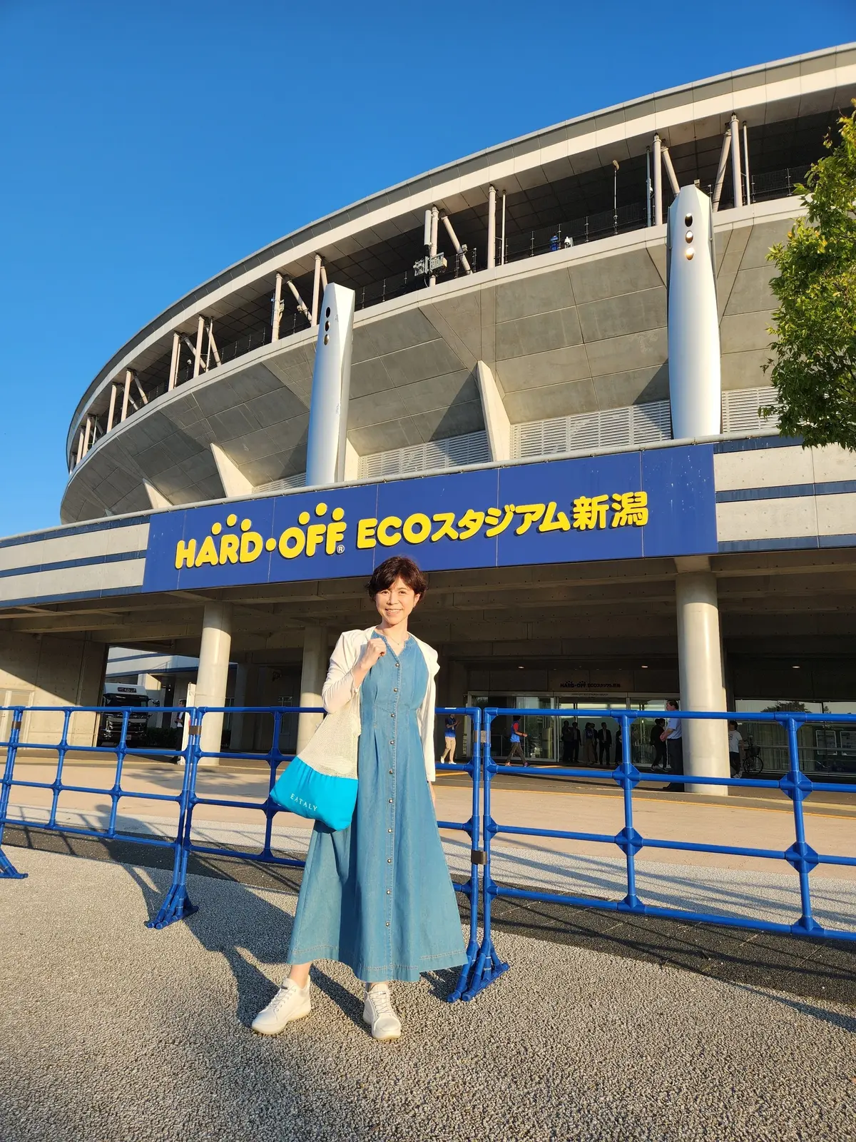 女性が青い長袖ワンピースで、ハードオフエコスタジアム新潟の入口前でポーズしています。手には青いトートバッグを下げ、白いスニーカーを履いています。背景にはコンクリート構造の建物と青いフェンスが並びます。