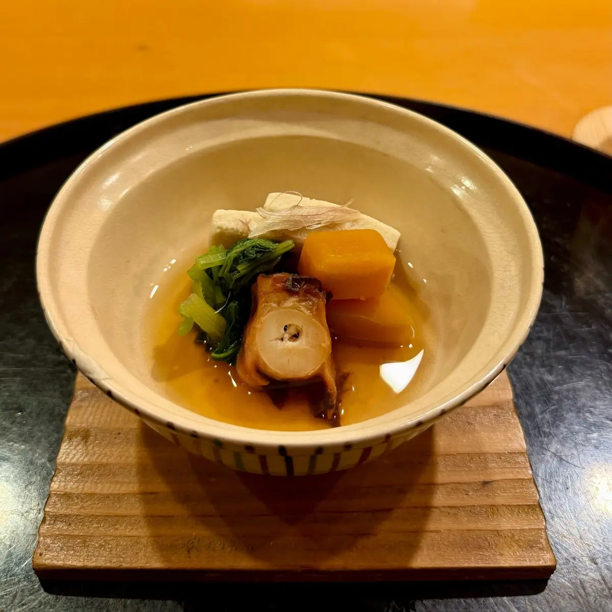 茶色の陶器鉢に盛り付けられた、和食の煮物。鶏肉、里芋、人参、小松菜が、薄い琥珀色の出汁で煮込まれている。木製の台に置かれ、背景はオレンジ色。