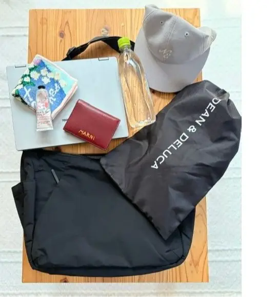 木製のテーブルの上に、ノートパソコン、ハンカチ、リップクリーム、財布、水筒、帽子、バッグ、布製のバッグが置かれている様子。バッグには「Dean & DeLuca」のロゴがプリントされている。