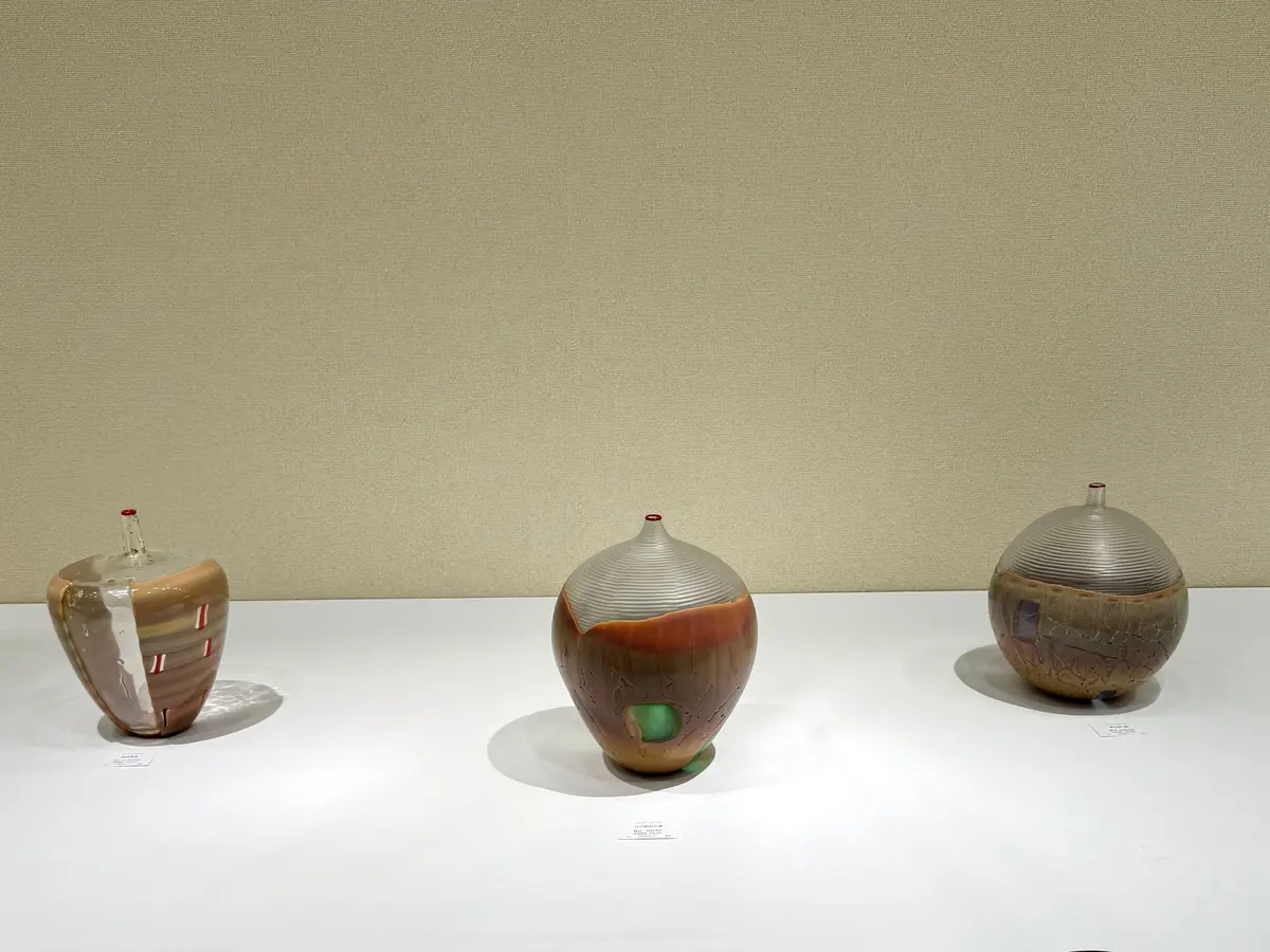 白い展示台に並んだ3つのガラス製のオブジェ。左は縦長の形状で赤と白のストライプ、中央は丸みを帯びた形状で茶色と緑の模様、右は球形に近い形状で茶色と紫の模様が施されている。
